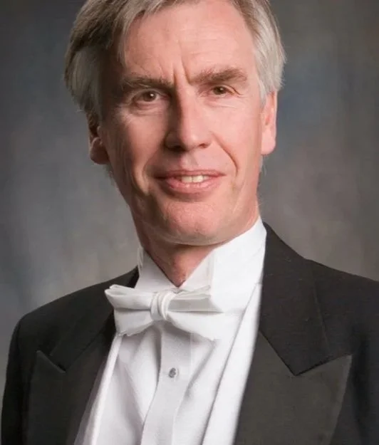 A man in formal tuxedo and white bow tie posing for a portrait against a neutral background.