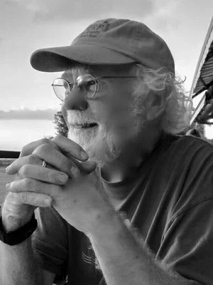 A smiling man with glasses, a cap, and curly hair, outdoors in black and white.