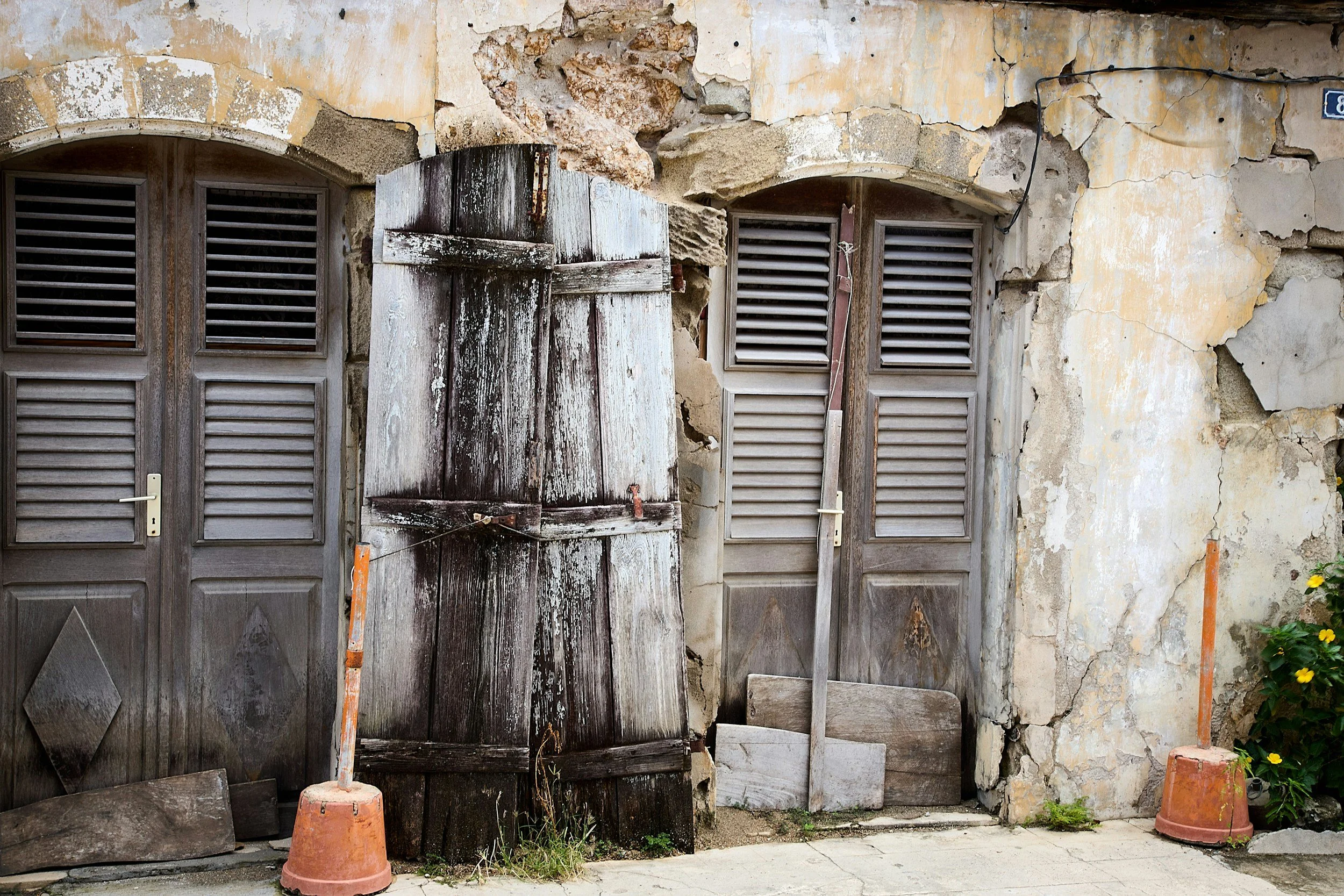 A dilapidated building facade with peeling paint and cracked plaster, featuring two closed wooden shutters covering windows, a weathered wooden door, construction cones, and a small green plant with yellow flowers in the corner.