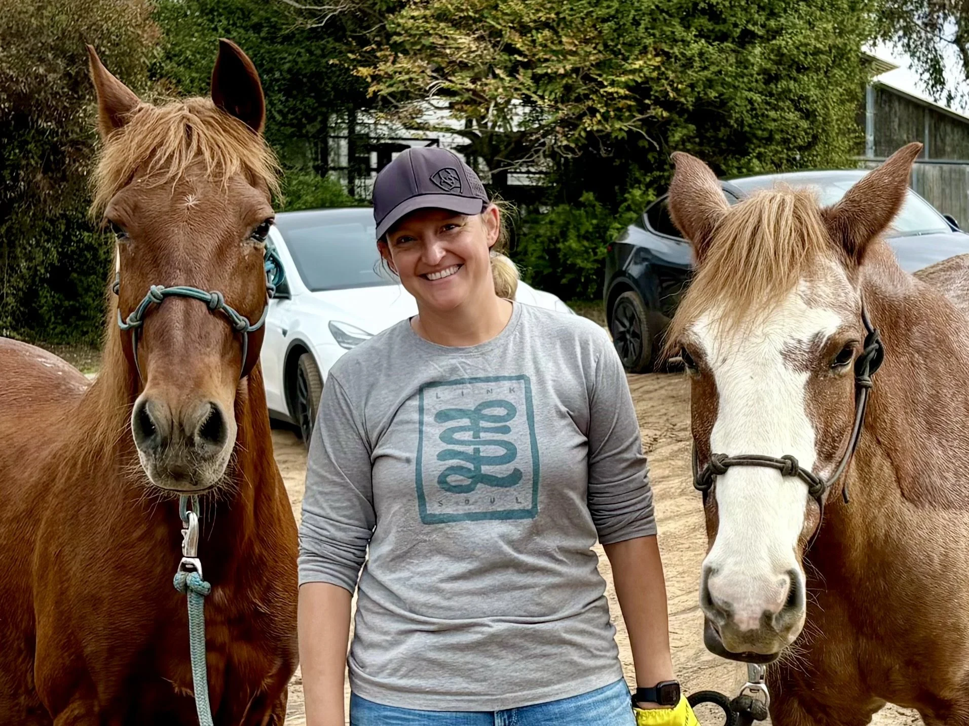 A woman smiling, wearing a gray long sleeve shirt and a baseball cap, standing between two horses outdoors.