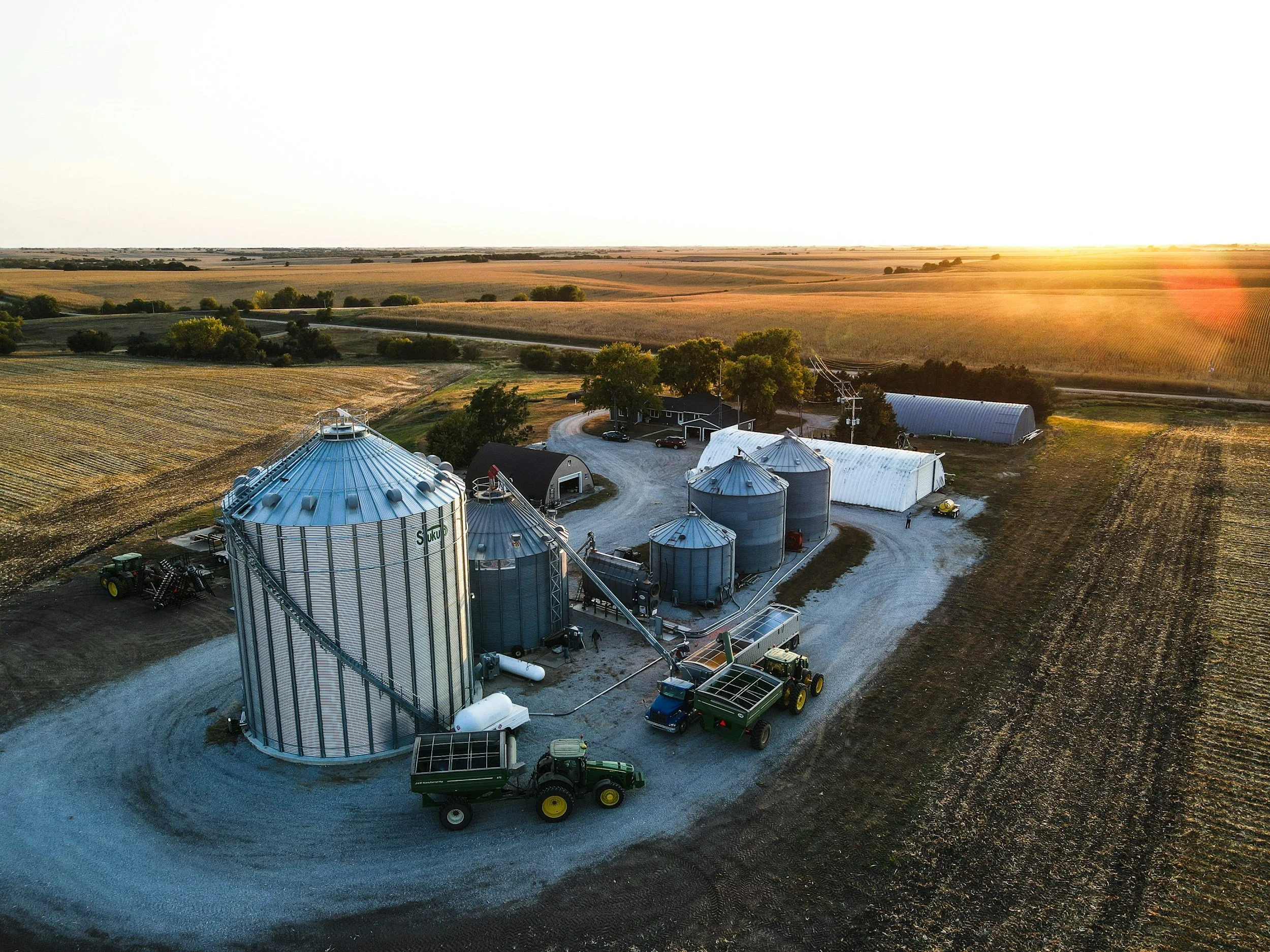Aerial view of a farm with silos, farm equipment, and several buildings, surrounded by fields and open land at sunset.