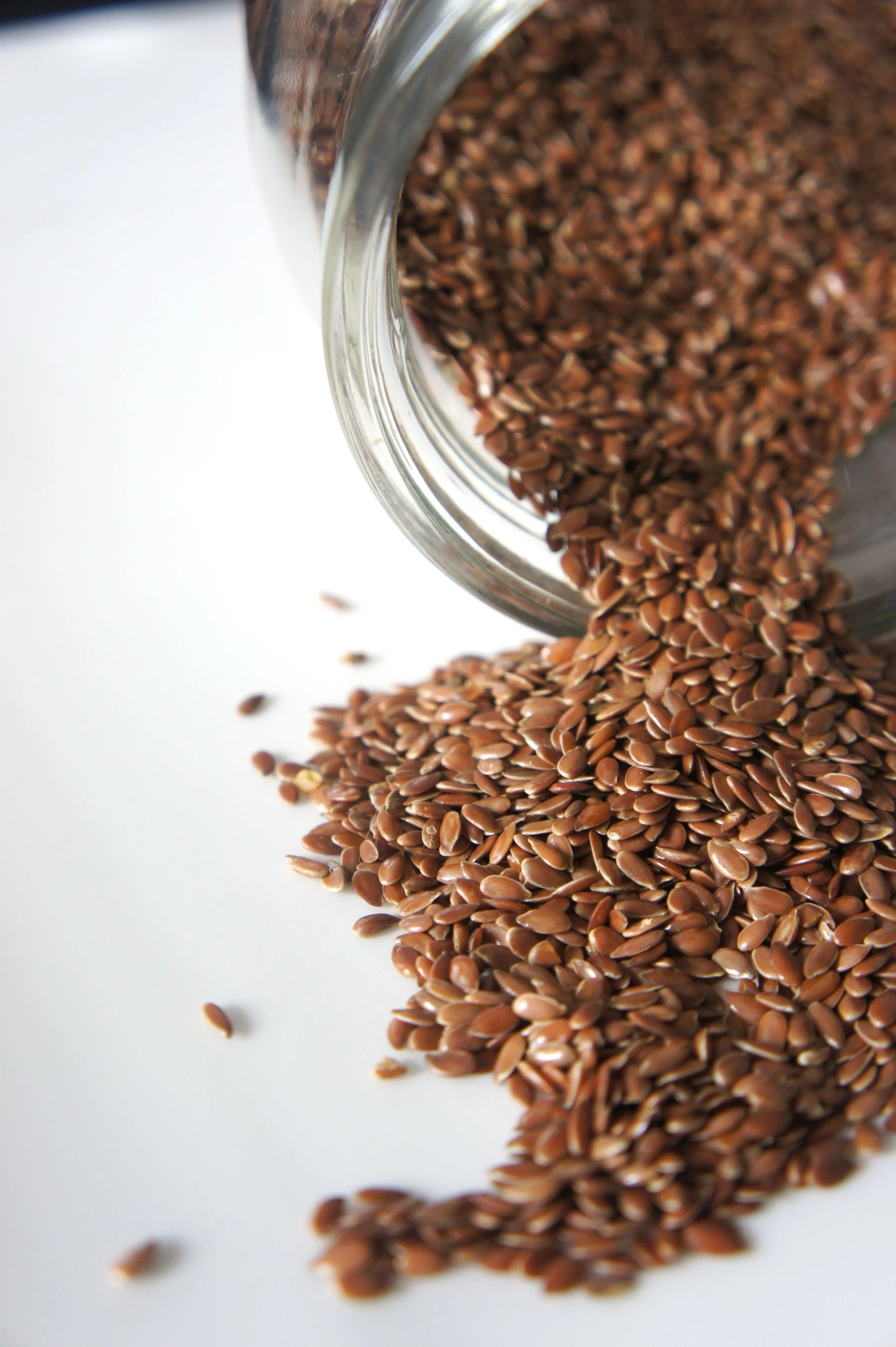 Flax seeds spilling out of a glass jar onto a white surface.