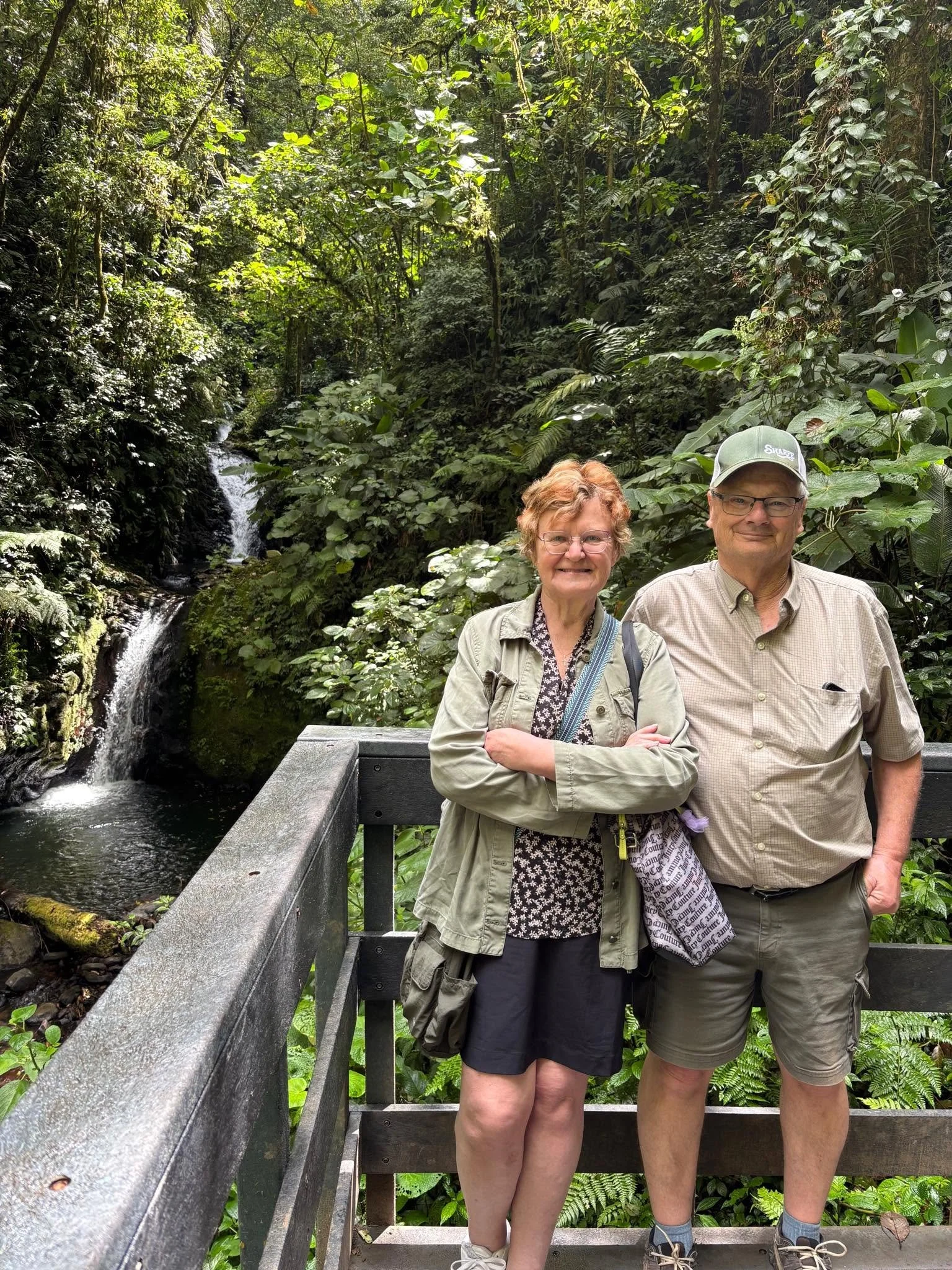 A smiling woman and a man stand on a bridge in a lush, green rainforest with a small waterfall in the background.