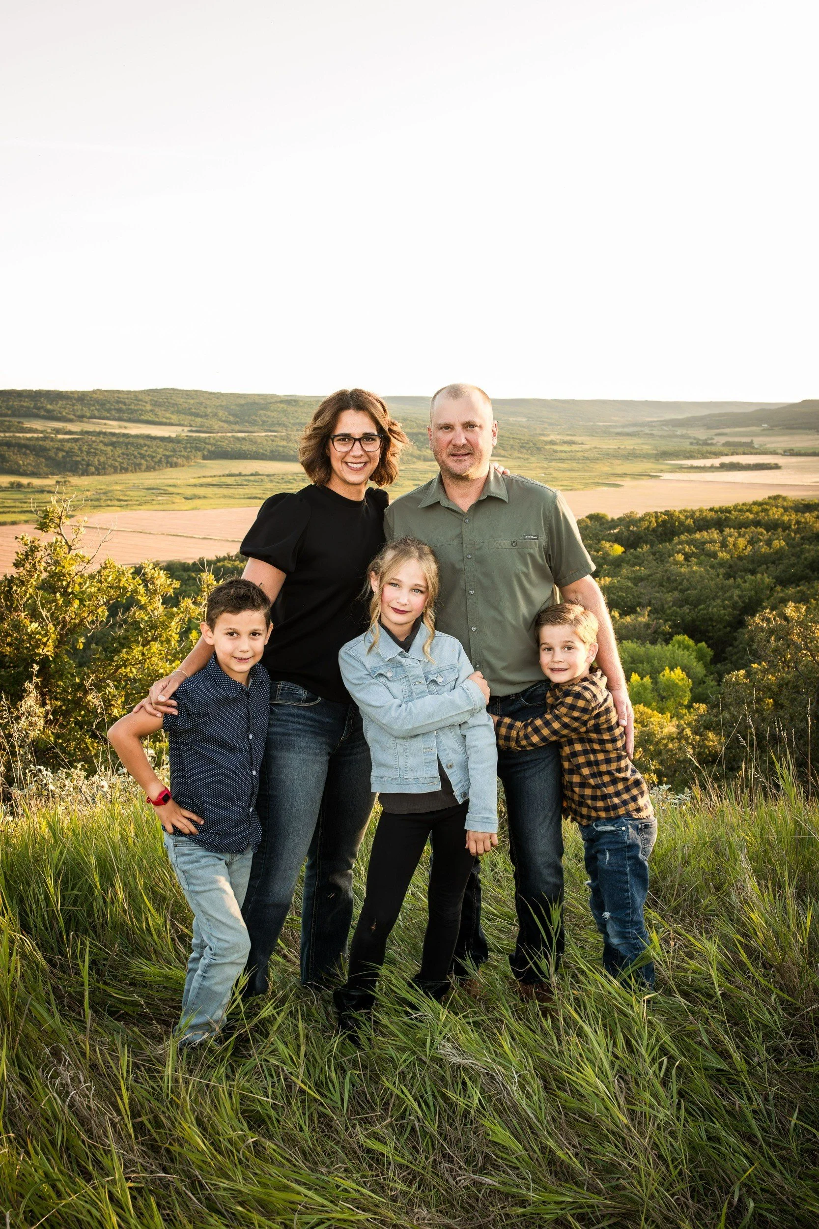 A family of six people standing outdoors in a grassy field with a landscape of rolling hills and farmland in the background during sunset. The group includes two adults and four children, smiling at the camera.