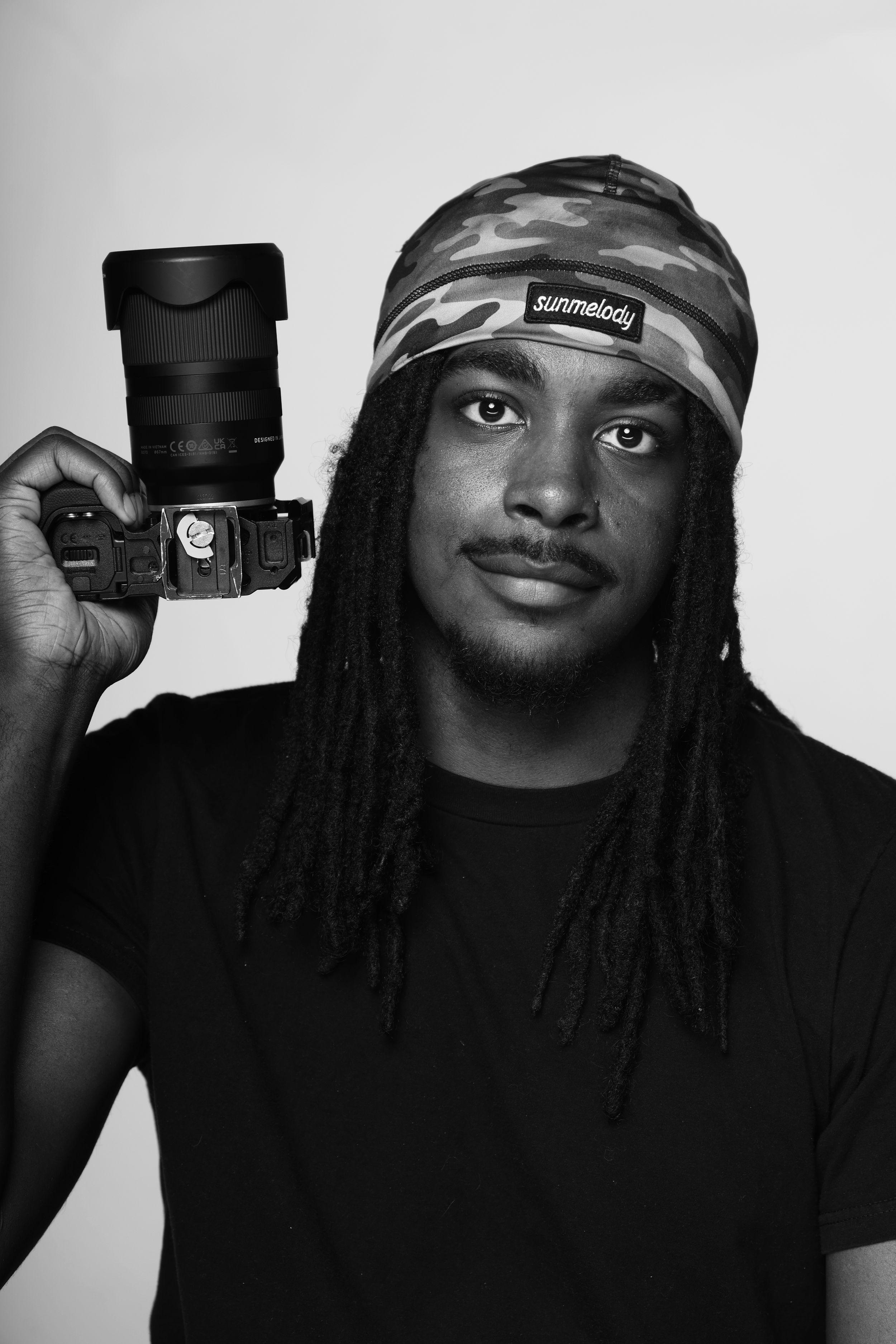 Black and white photo of a young man with long dreadlocks, wearing a camouflage headscarf labeled 'sunmelody,' holding a camera near his face.