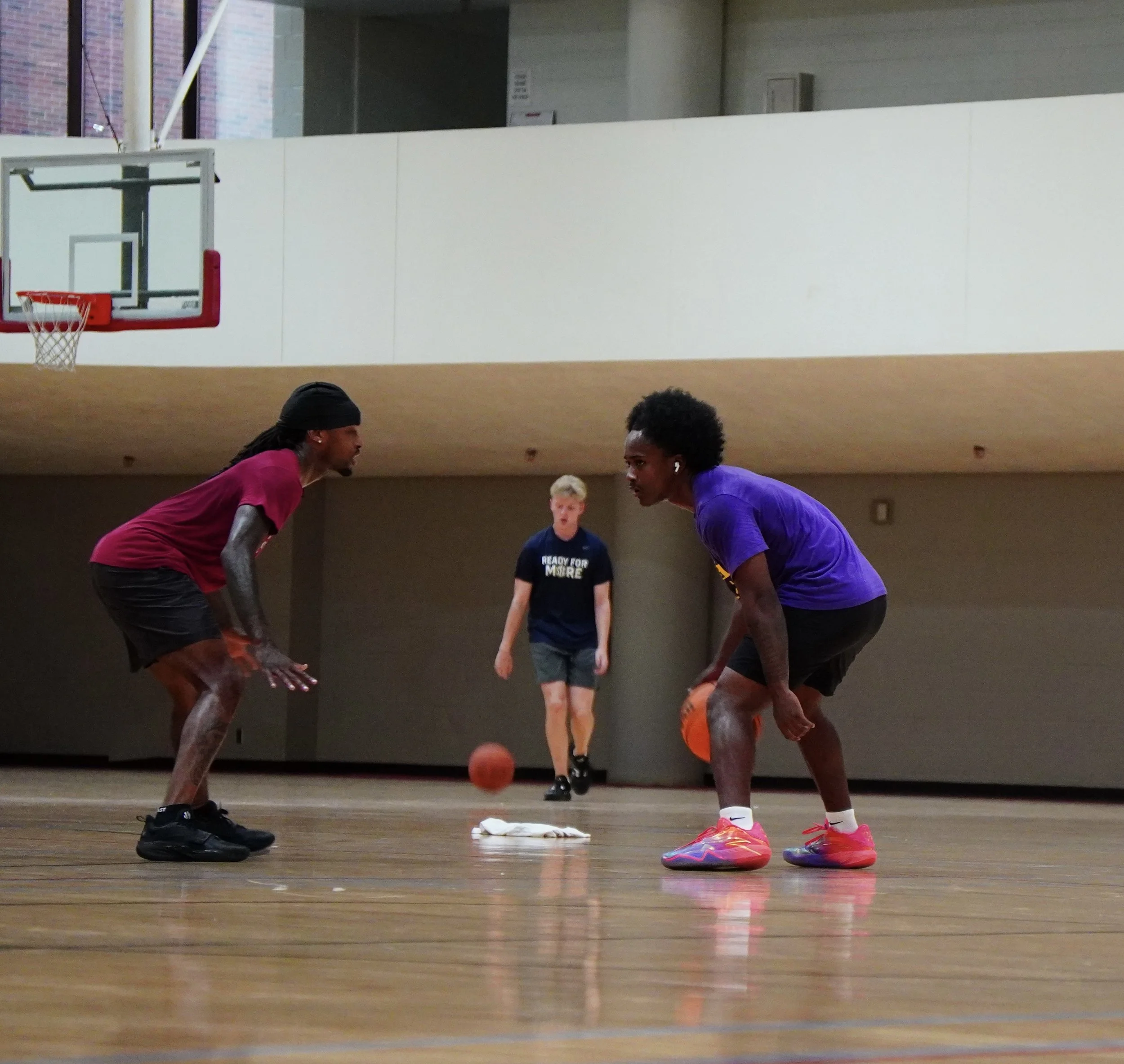 Two young men in basketball practice or game, crouching near a basketball on a wooden court, with a coach or referee observing in the background.