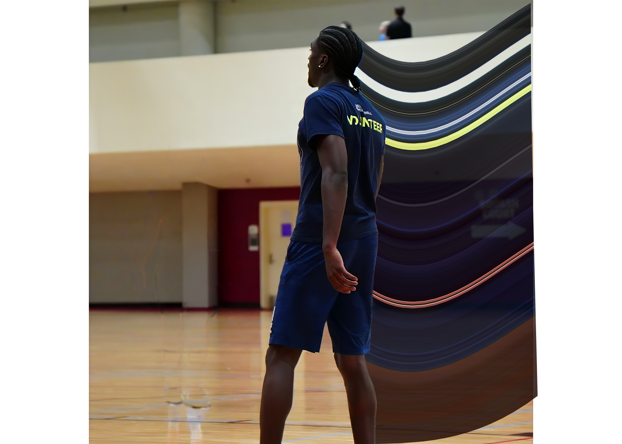 A person standing in a gymnasium wearing a blue sports T-shirt and shorts, with braided hair, appears to be a volunteer for some event.