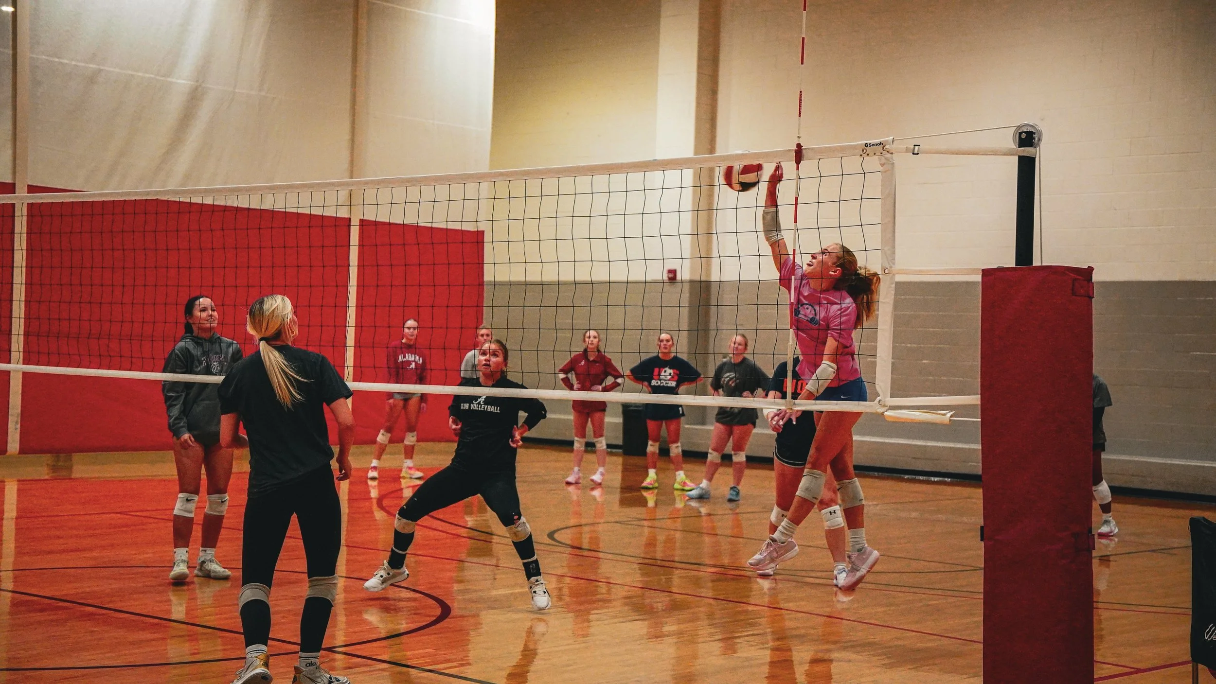 Women playing volleyball indoors with one jumping to block the ball at the net, several other players watching and preparing, on a gymnasium court.