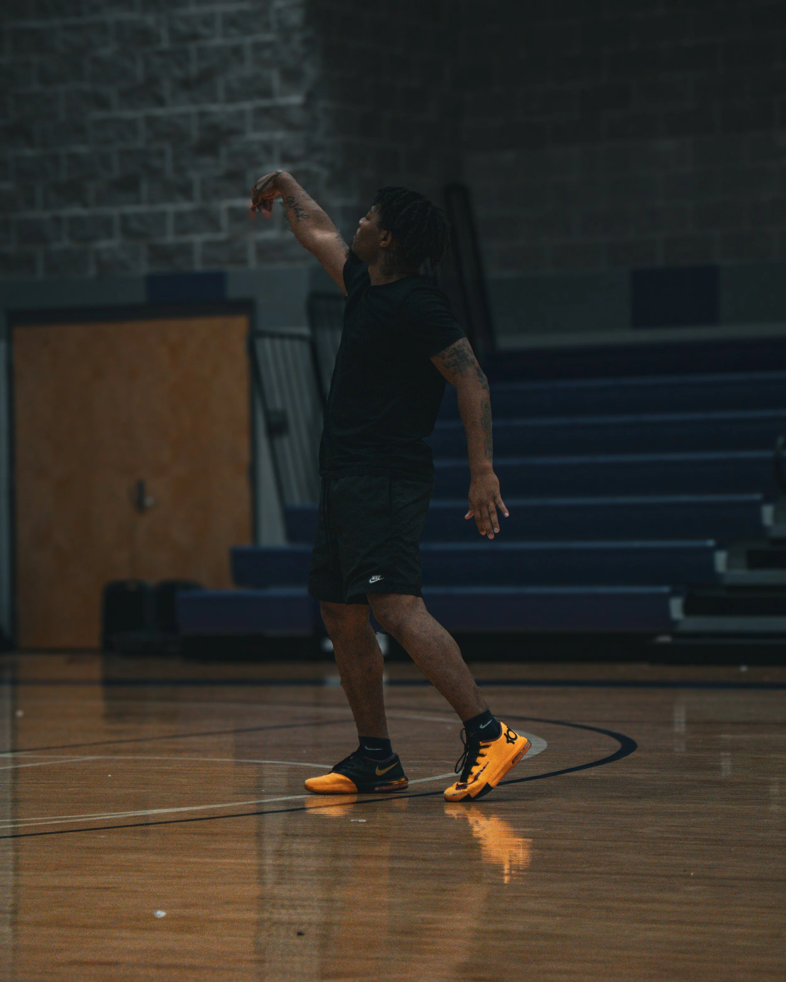 A person with dreadlocks and tattoos practicing dance or movement in a gymnasium, wearing a black t-shirt, black shorts, and yellow-orange Nike sneakers.