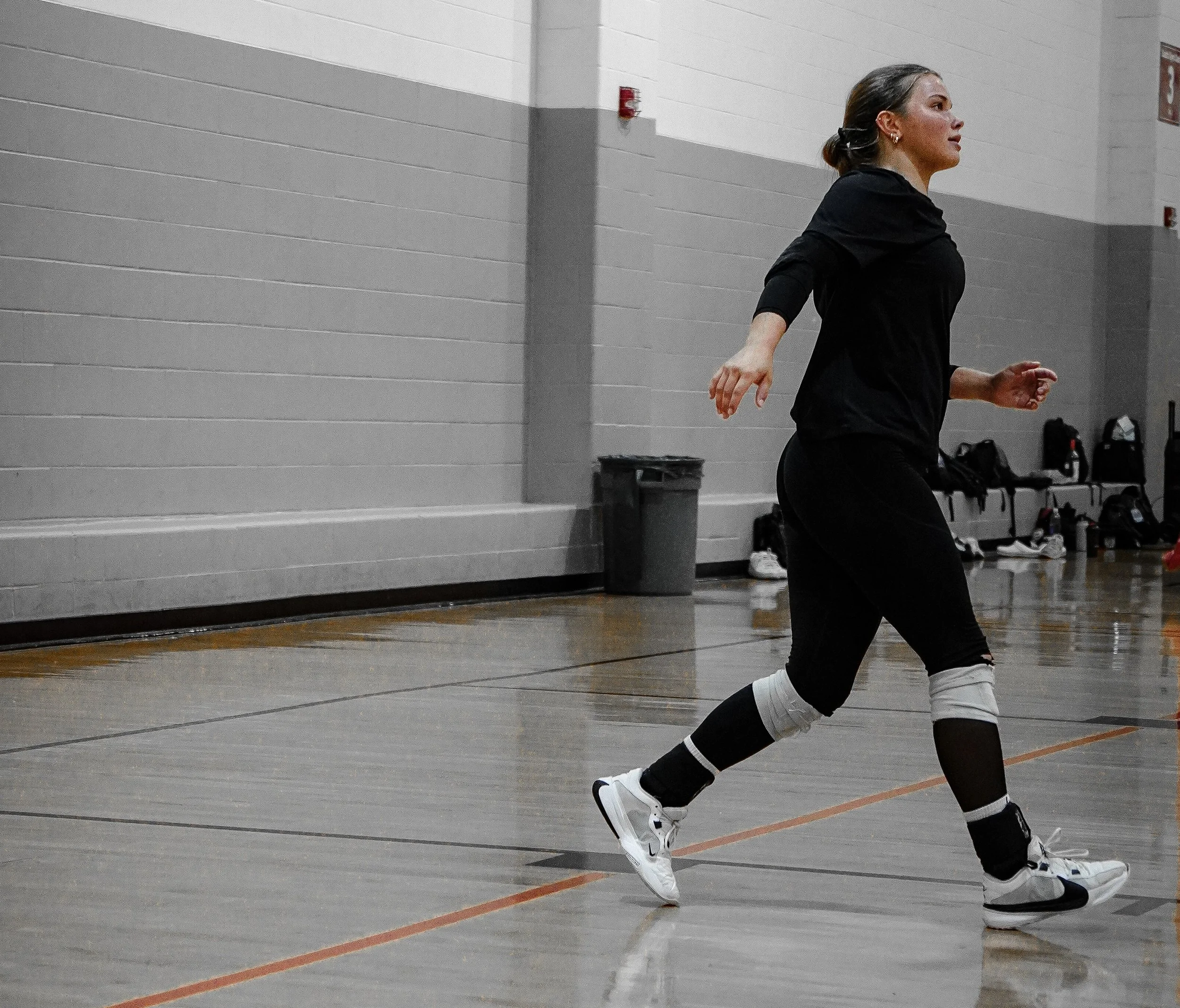 A woman dressed in black athletic clothing and white sneakers is running or jogging inside a gymnasium or sports hall with beige walls and a hardwood floor.