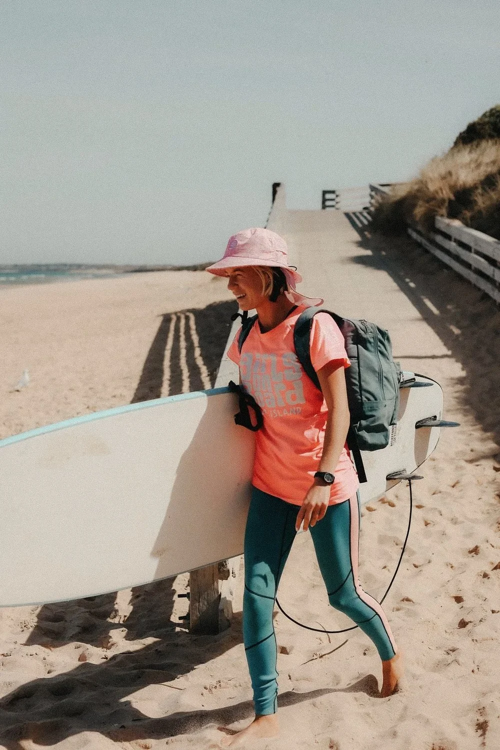 A young woman with a pink hat, pink t-shirt, and teal leggings carrying a backpack and holding a surfboard walking on a sandy beach near a boardwalk with stairs.