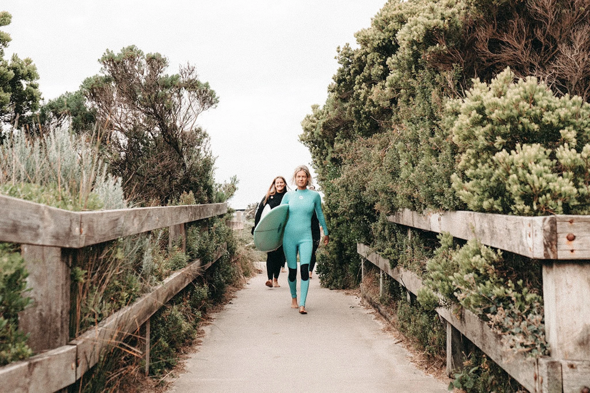 A woman in a turquoise wetsuit carrying a surfboard walking on a wooden bridge surrounded by dense green bushes and trees.