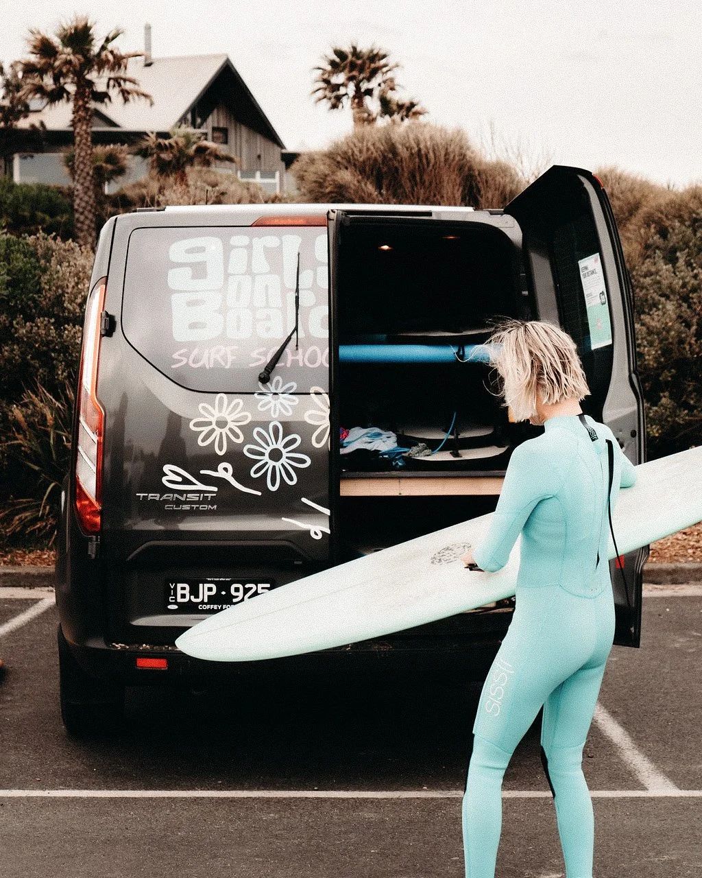 A woman in a light blue wetsuit holding a surfboard, standing near an open surf shop van with surf gear inside, in a parking lot with trees and houses in the background.