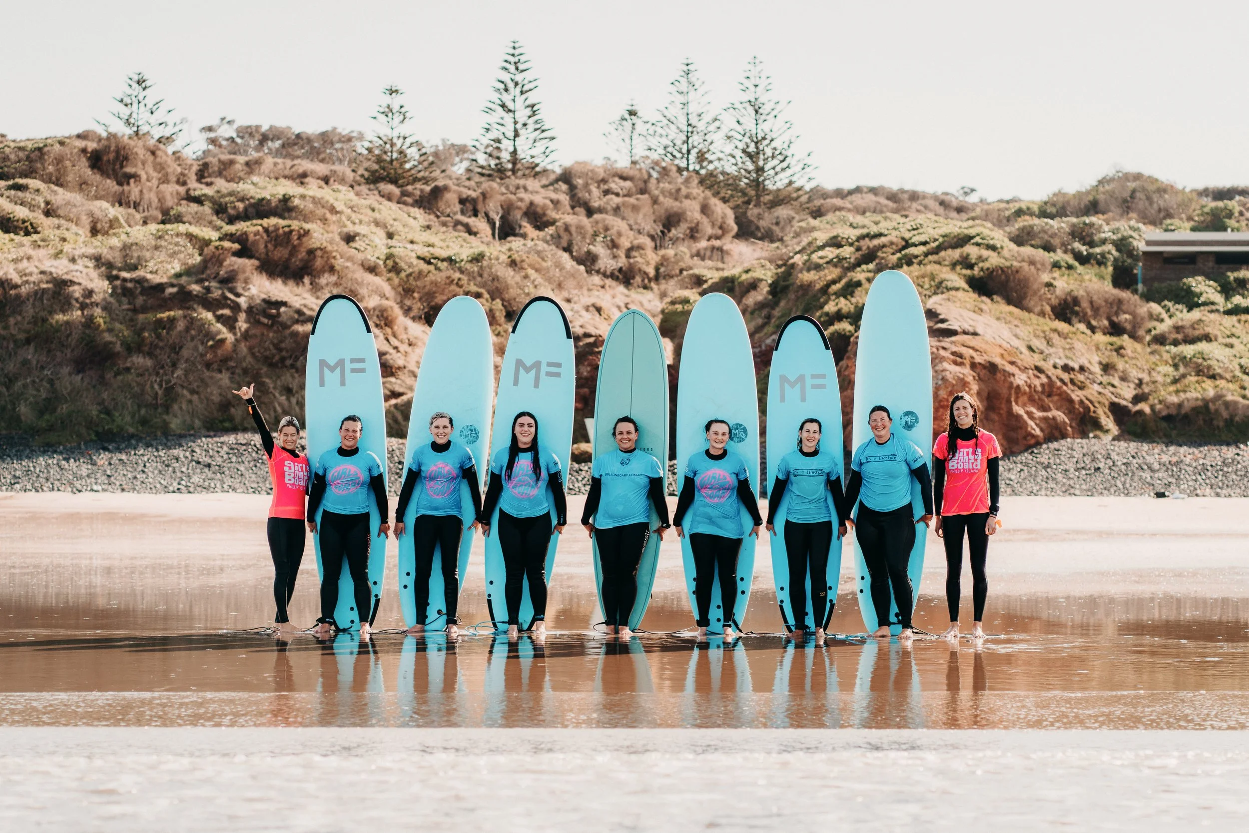 A group of women in matching blue and pink shirts holding surfboards stands on a beach with a rocky and wooded hillside in the background.