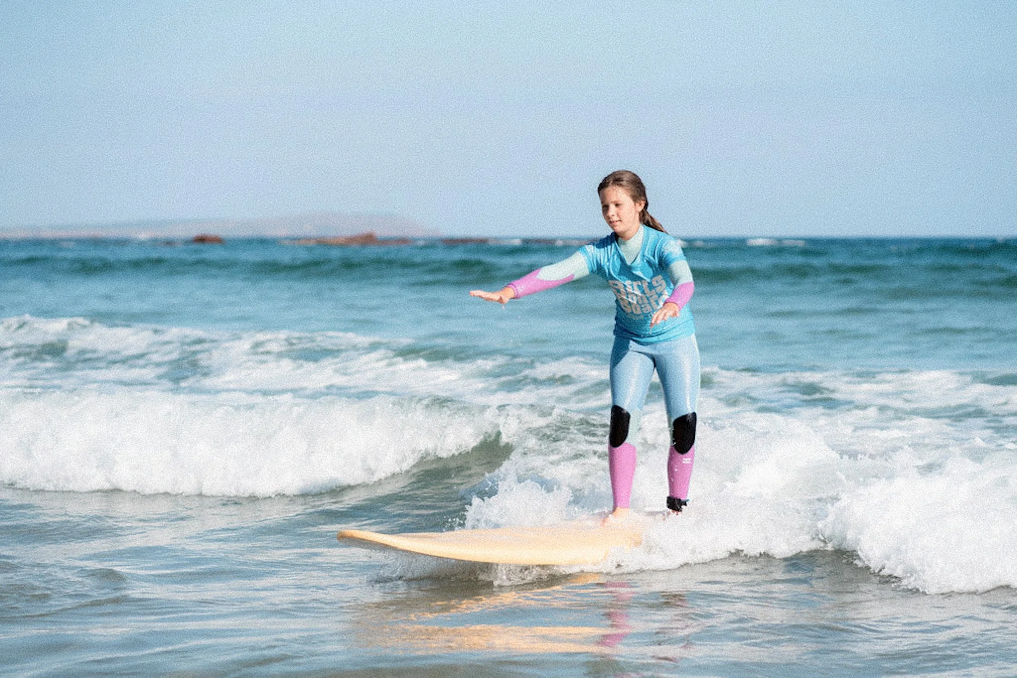 A person in a blue wetsuit and pink flotation gear balancing on a surfboard in the ocean.