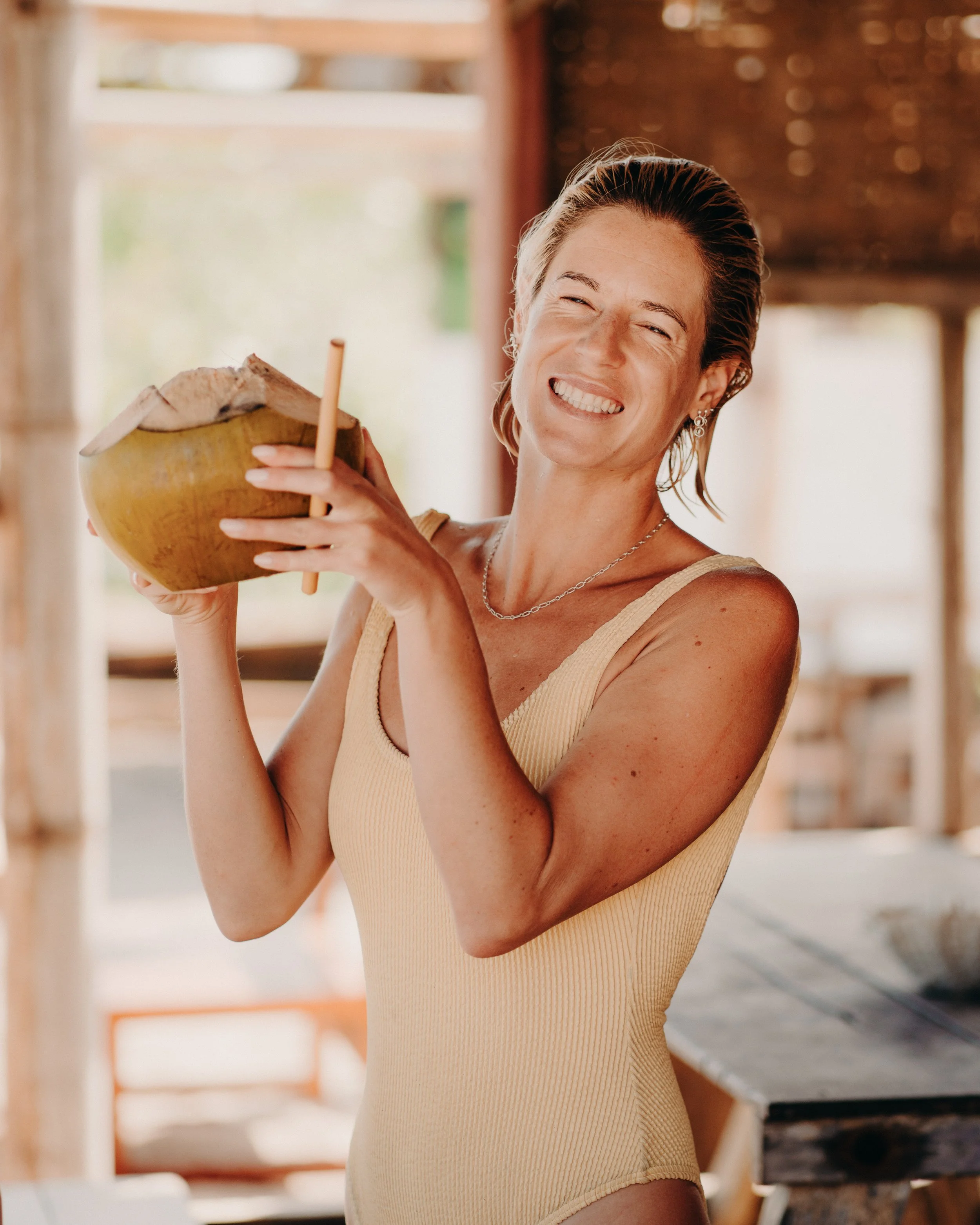 A smiling woman with short hair and earrings holding a coconut drink with a straw indoors with warm lighting and wooden furniture.