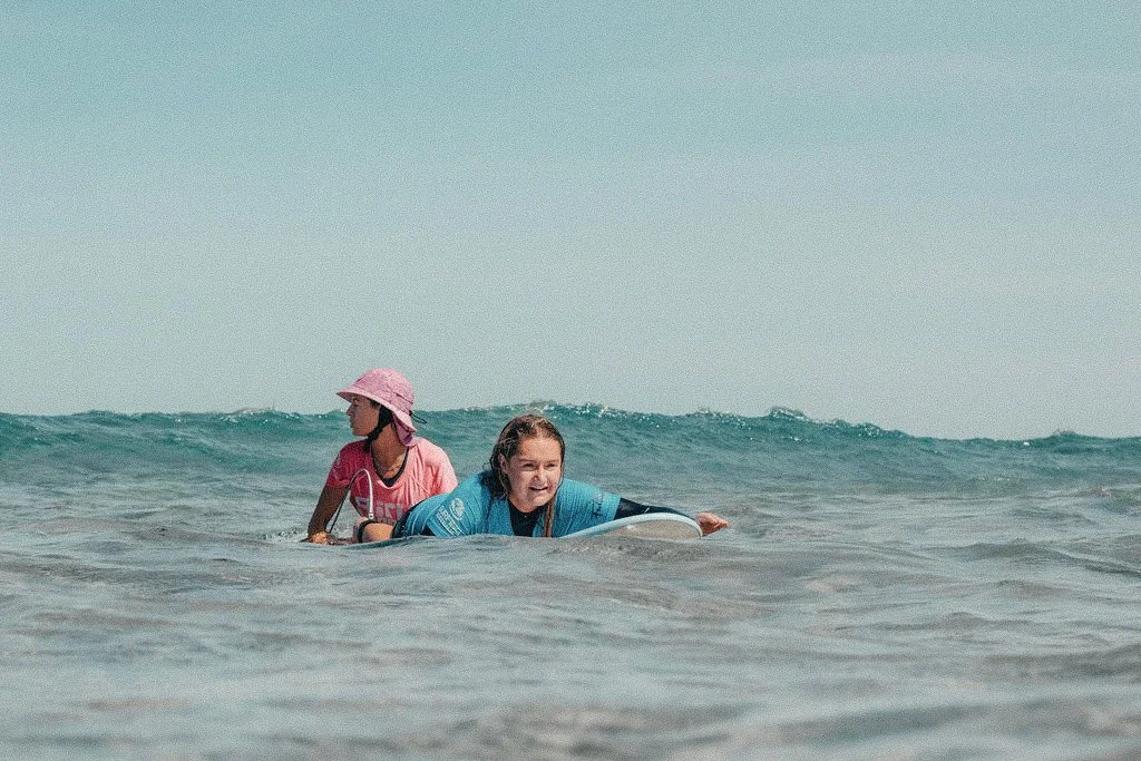 Two children in the ocean, with one lying on a surfboard and the other sitting in the water, both wearing swimwear and sun hats.