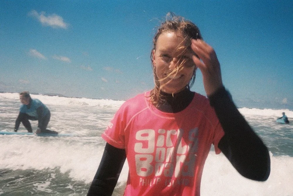 A woman in a pink t-shirt and black long sleeves smiling at the camera while standing in the ocean with waves, with two other people surfing in the background under a partly cloudy sky.