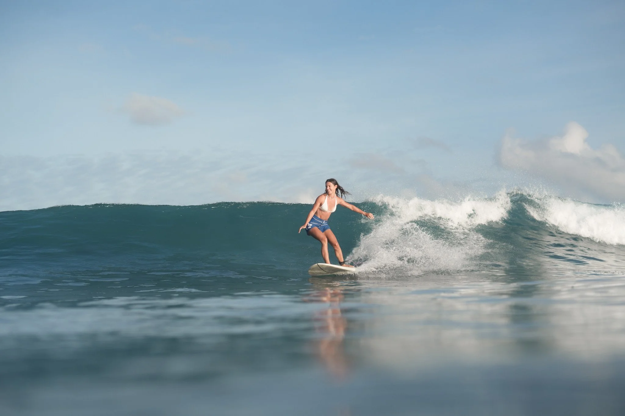 A young woman in a white bikini top and blue shorts surfing on a wave under a partly cloudy sky.