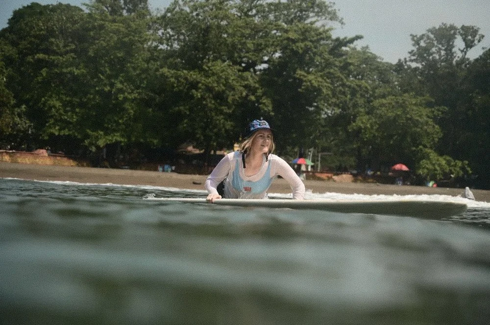 A woman lies on a surfboard in the water near a sandy beach, smiling, with trees and umbrellas in the background.