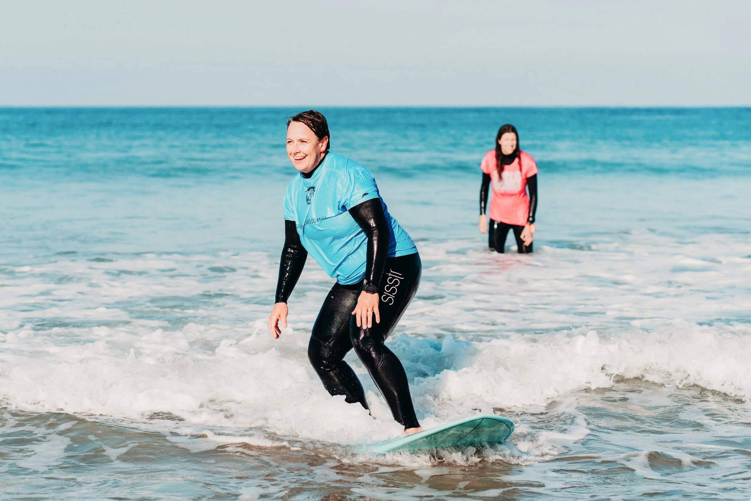 Two women surfing in the ocean, one in a blue shirt and black wetsuit, smiling, with another woman in a pink shirt in the background.