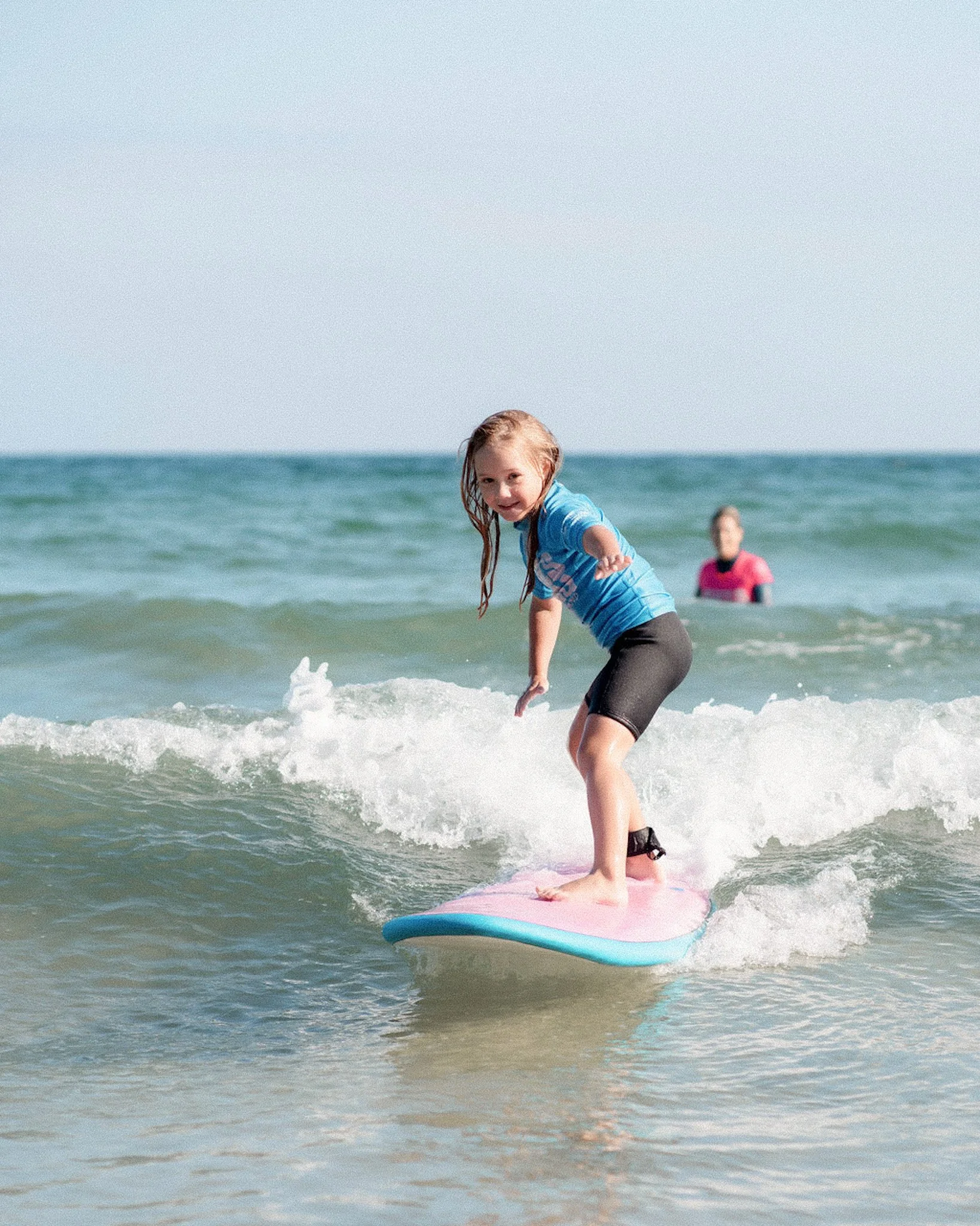 A young girl with wet hair surfing on a pink surfboard in the ocean, with another person in a pink shirt in the background.