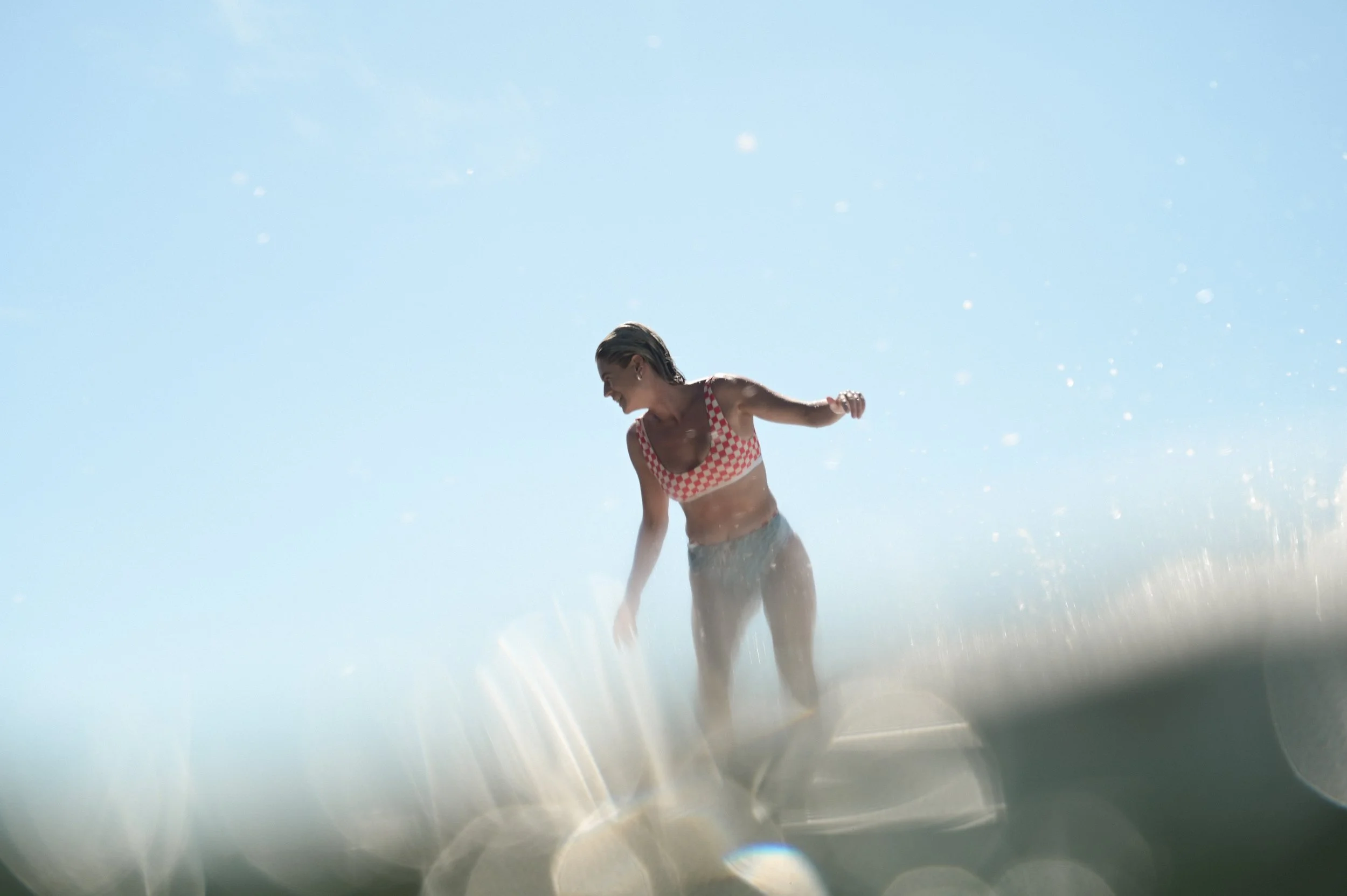 A woman in a checkered bikini is enjoying a moment at the beach, with sunlight and water droplets surrounding her.
