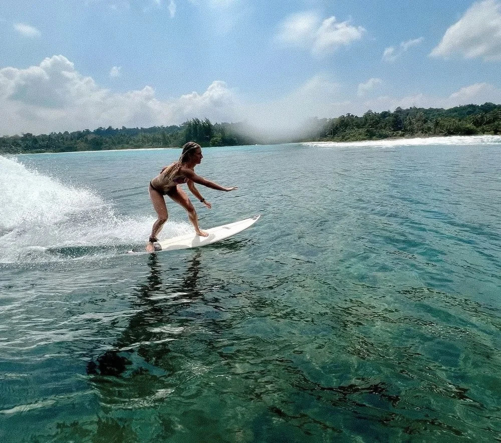 Woman surfing on a clear body of water under a partly cloudy sky, with a distant shoreline and trees in the background.
