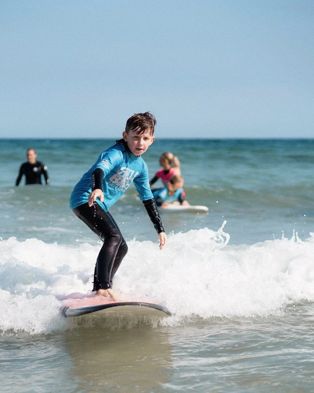 A young boy learning to surf on the ocean with other surfers in the background on a sandy beach.