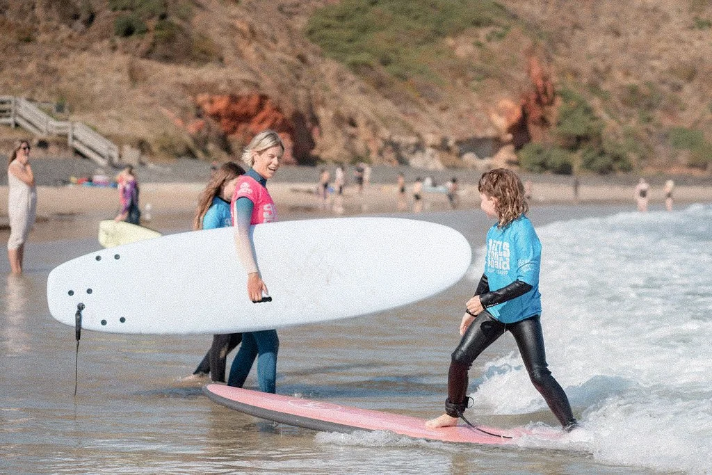 Two women at the beach, one holding a surfboard and the other standing on a surfboard, smiling and talking with others in the background.
