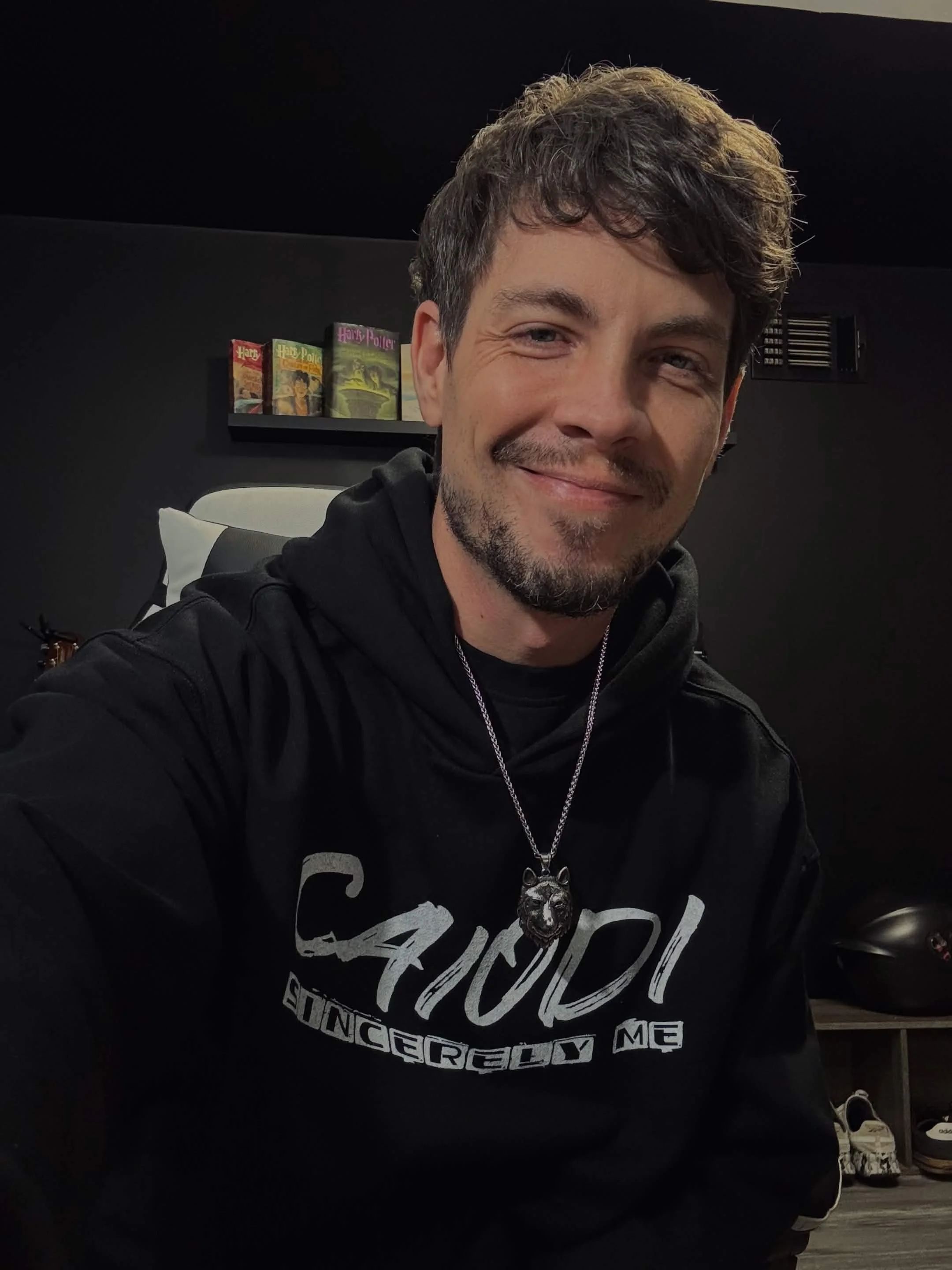 A young man with dark hair and a beard smiling for a selfie in a room with black walls. Behind him, a black shelf with several Harry Potter books. He's wearing a black hoodie with white text and a large necklace with a wolf head pendant.