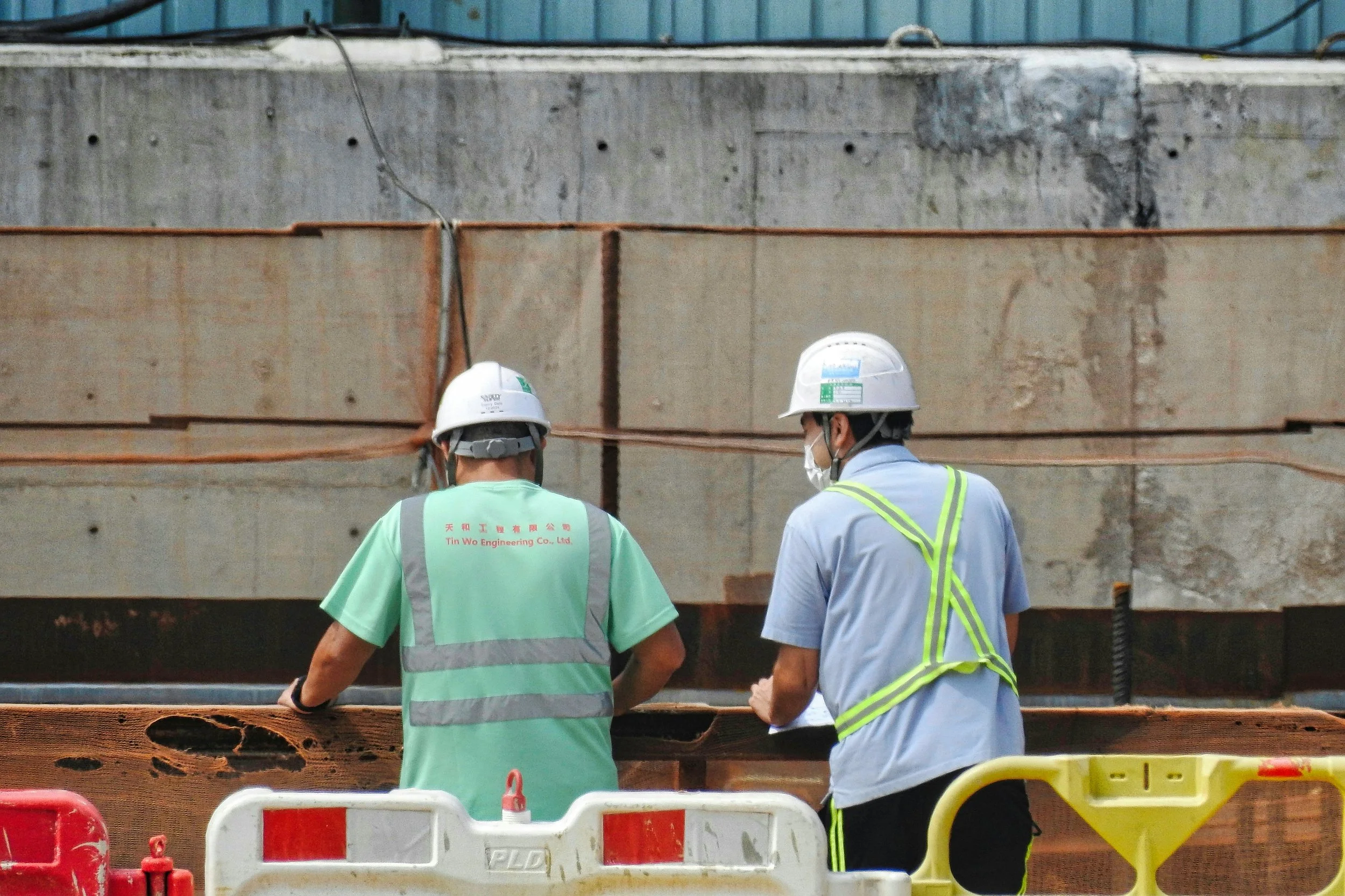 Two construction workers wearing helmets and safety vests standing near a construction site with large concrete structures and barriers.