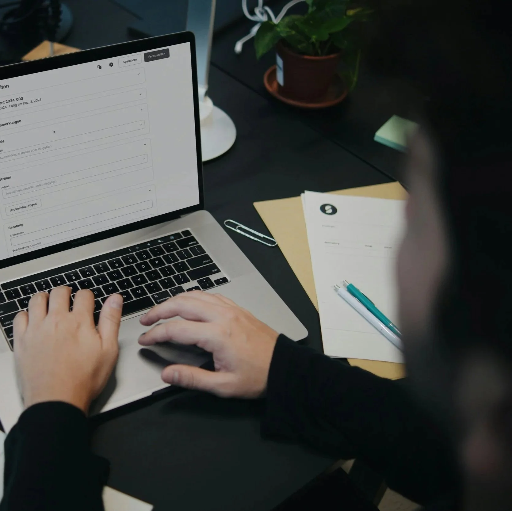 Person using a laptop with documents and pens on a black desk, potted plant and sticky notes in the background.