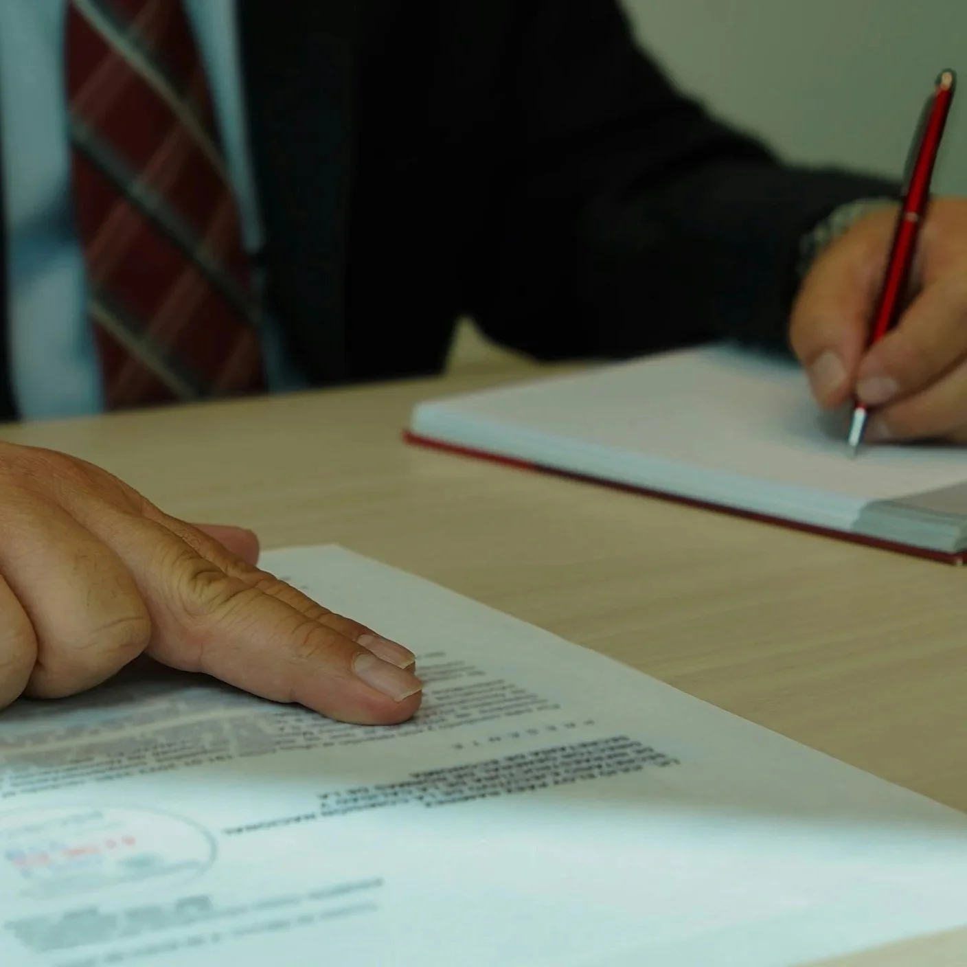 A person pointing at a document on a table while another person writing in a notebook.