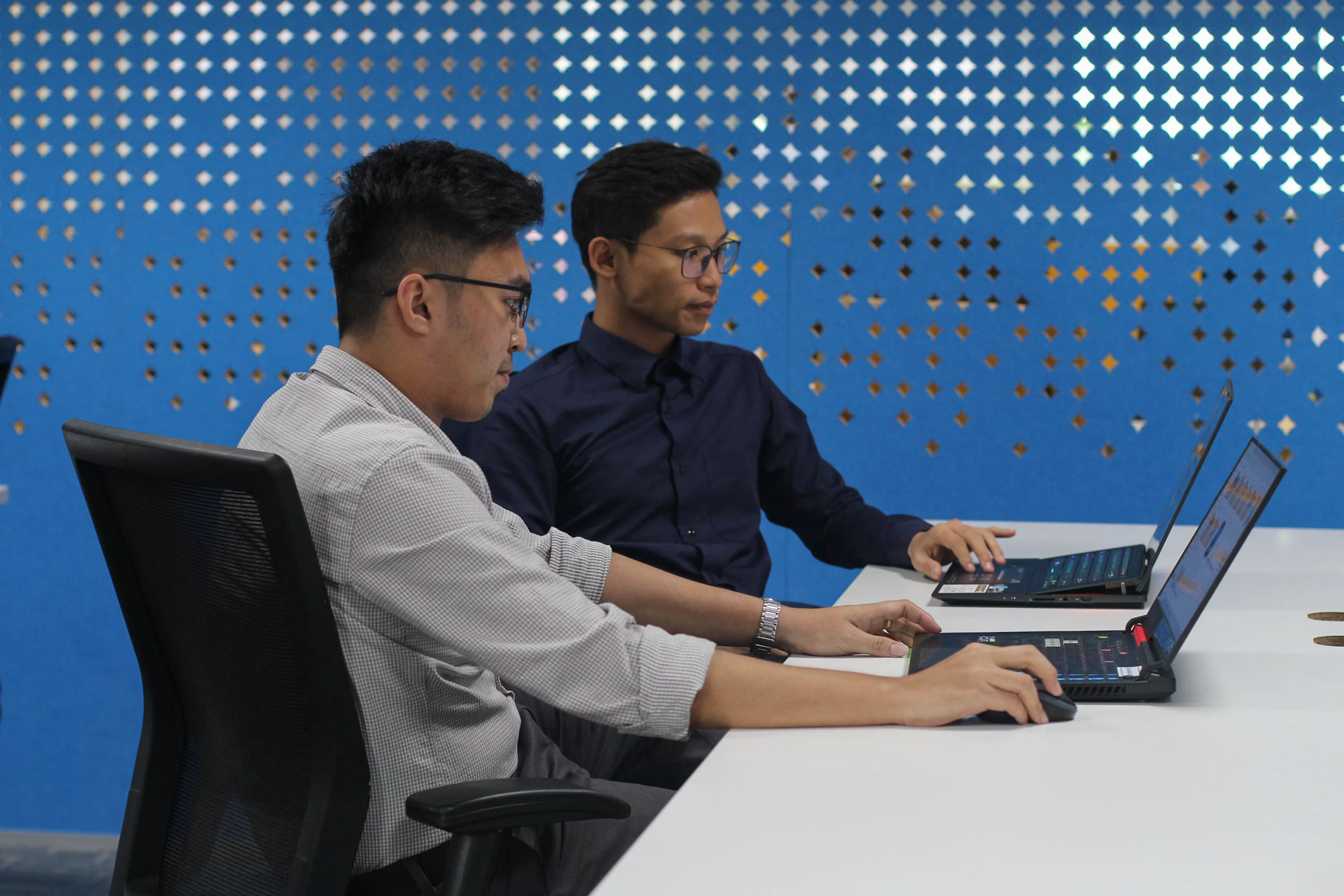 Two men sitting at a white table working on laptops in a conference room with a blue decorative wall.