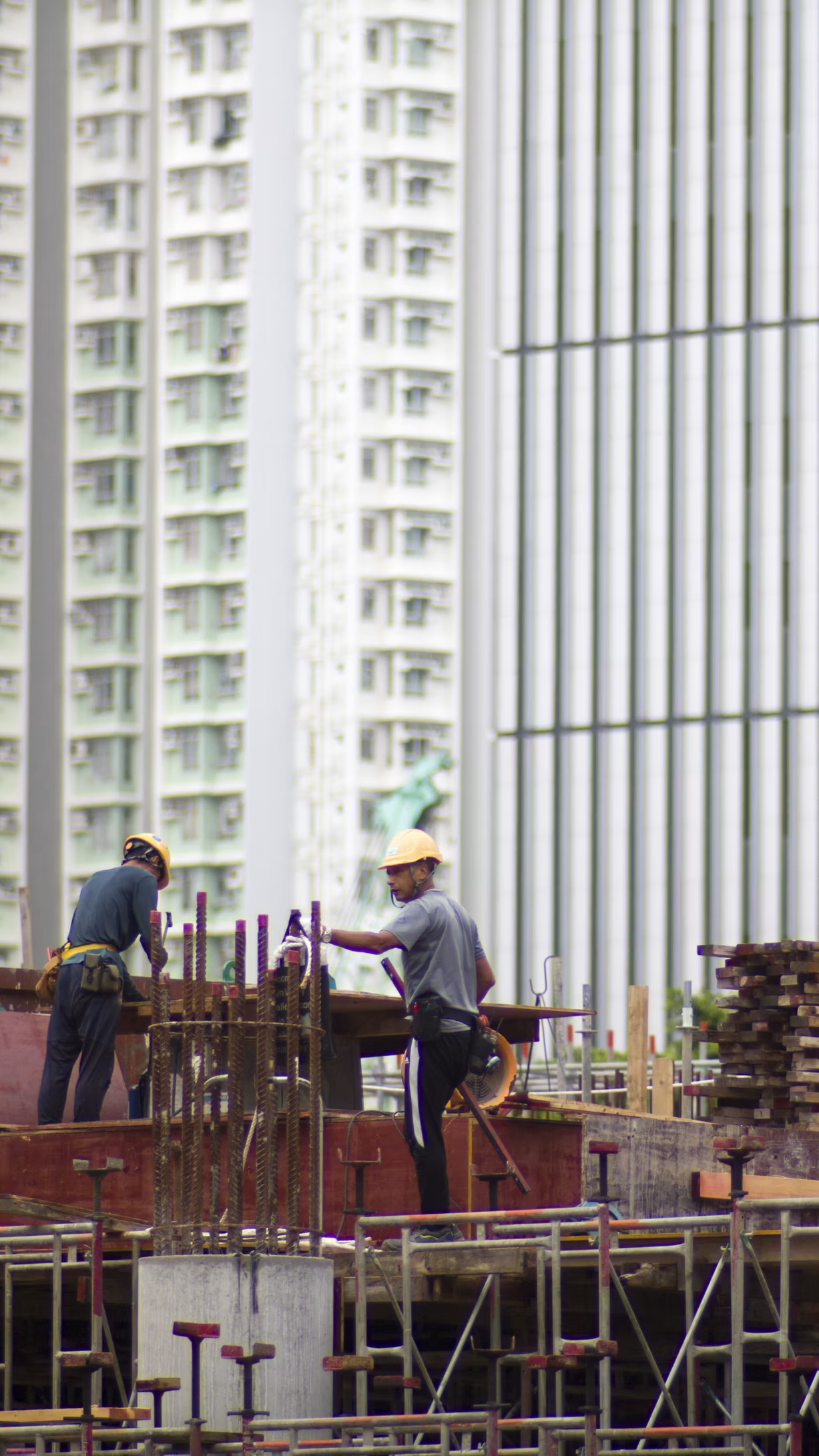 Construction workers wearing yellow helmets working on a building construction site with high-rise buildings in the background.