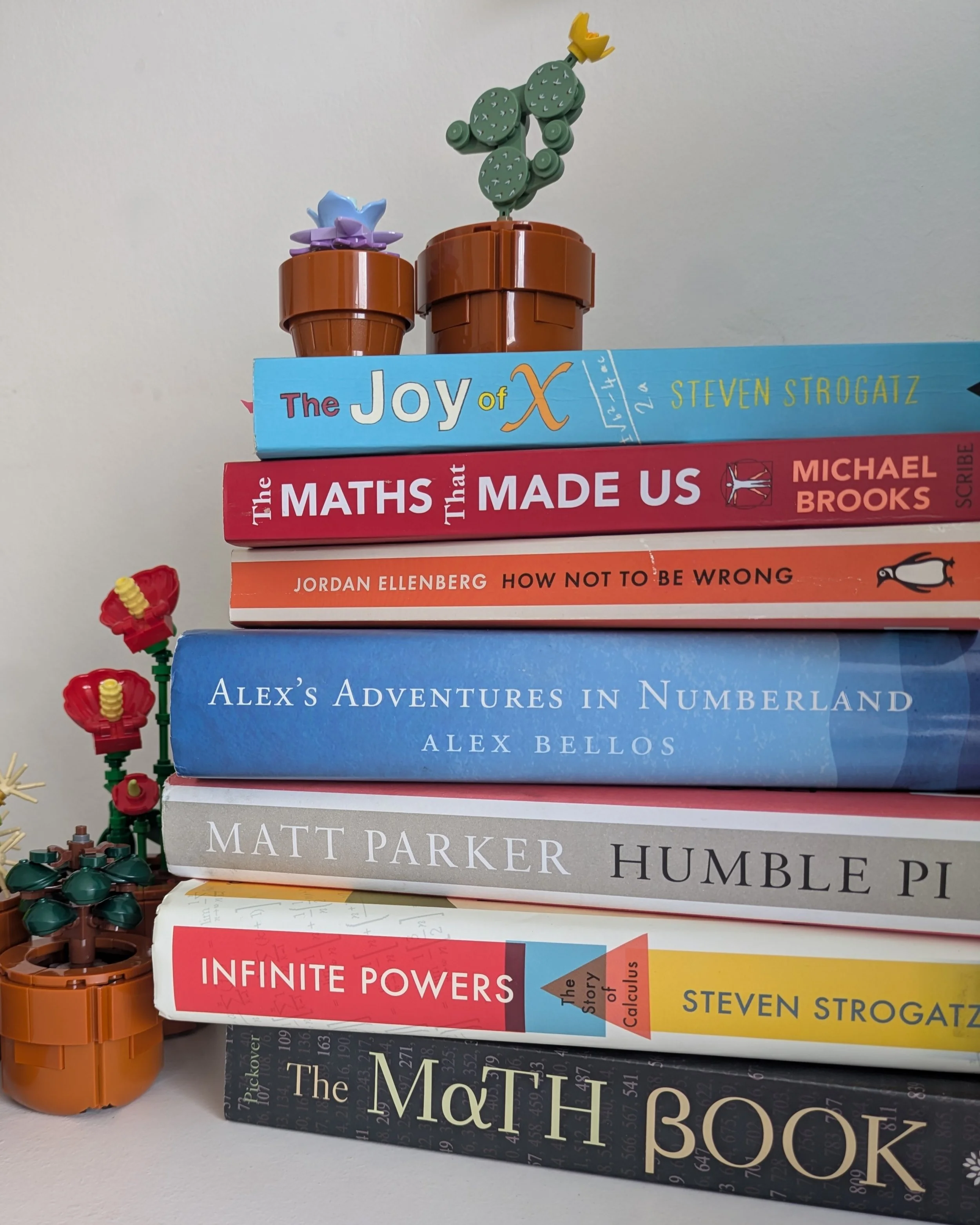 Stack of books on a white shelf, with a Lego cactus plant and a Lego flower in pots on top, against a plain wall.