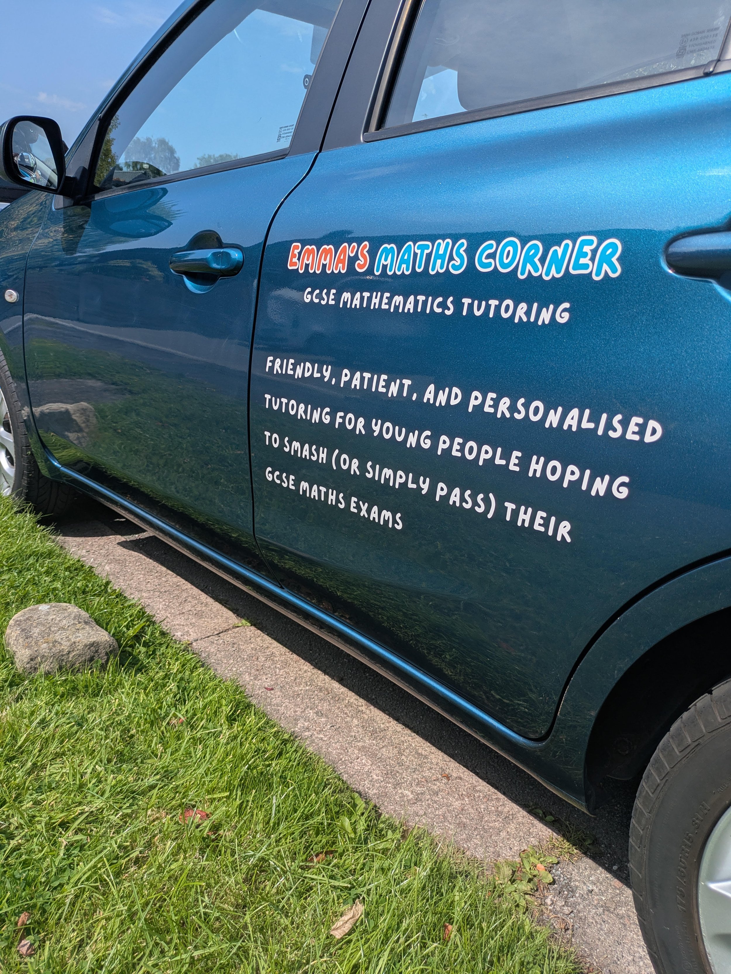 Blue car with sign on the side that reads 'Emma's Maths Corner', offering GCSE mathematics tutoring, parked on a grassy roadside.