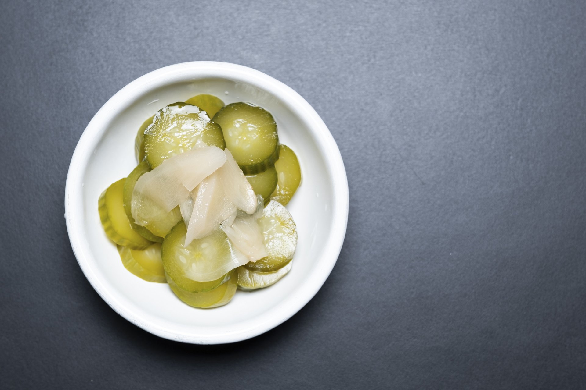 A small white bowl containing sliced pickles and onions on a dark gray surface.