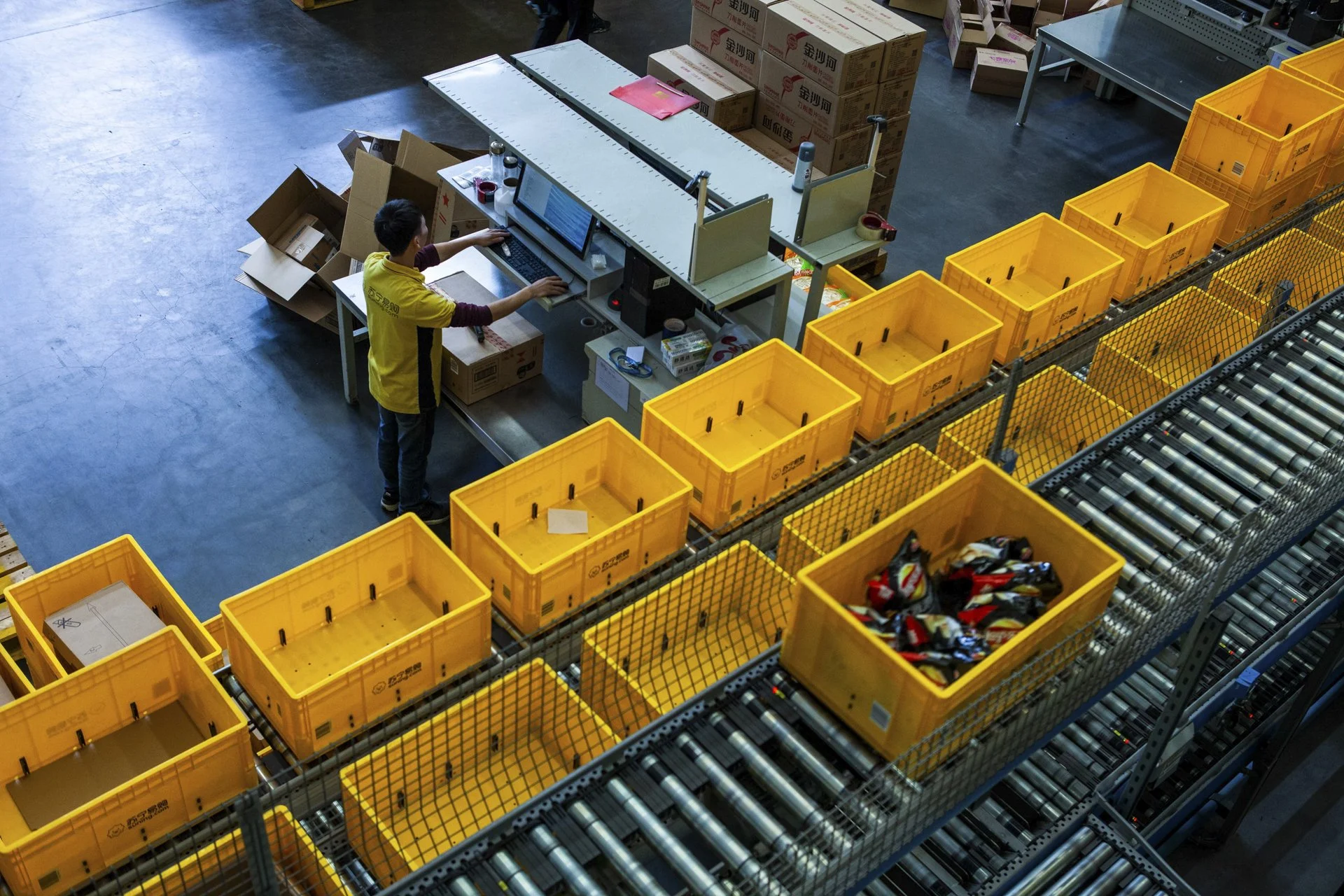 A warehouse worker at a conveyor belt with multiple yellow bins filled with sorted packages and products, surrounded by stacks of cartons and storage boxes.
