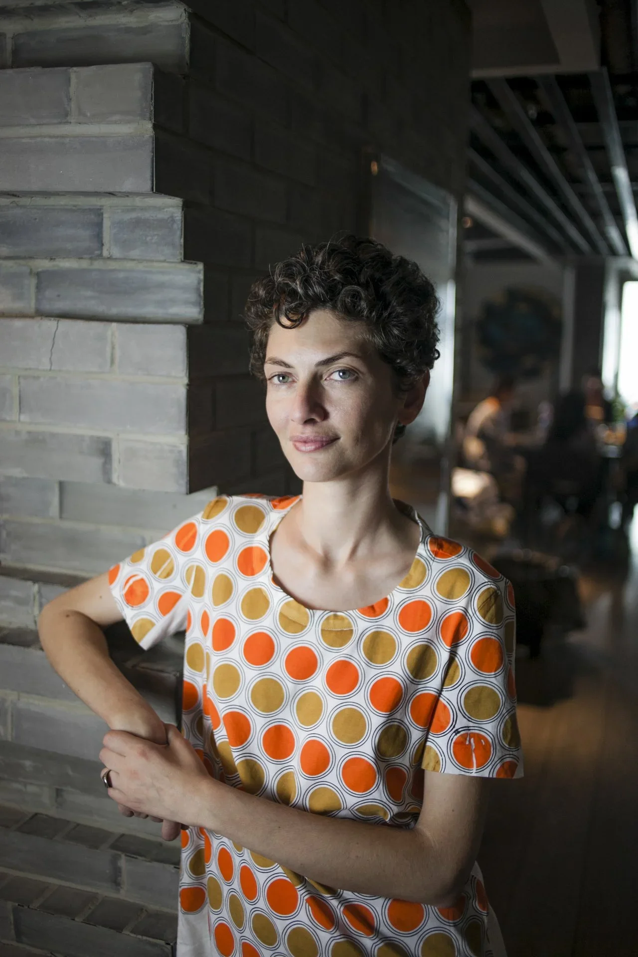 A woman with short, curly dark hair and blue eyes standing indoors next to a gray brick wall, wearing a white dress with a pattern of orange, yellow, and brown circles, smiling softly with her left hand on her waist.