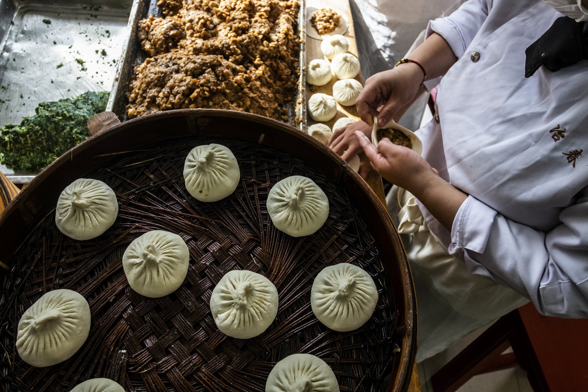 A person preparing steamed dumplings, with a bamboo tray of plain dumplings in the foreground and a tray of topped minced meat at the back, in a kitchen or food stall setting.