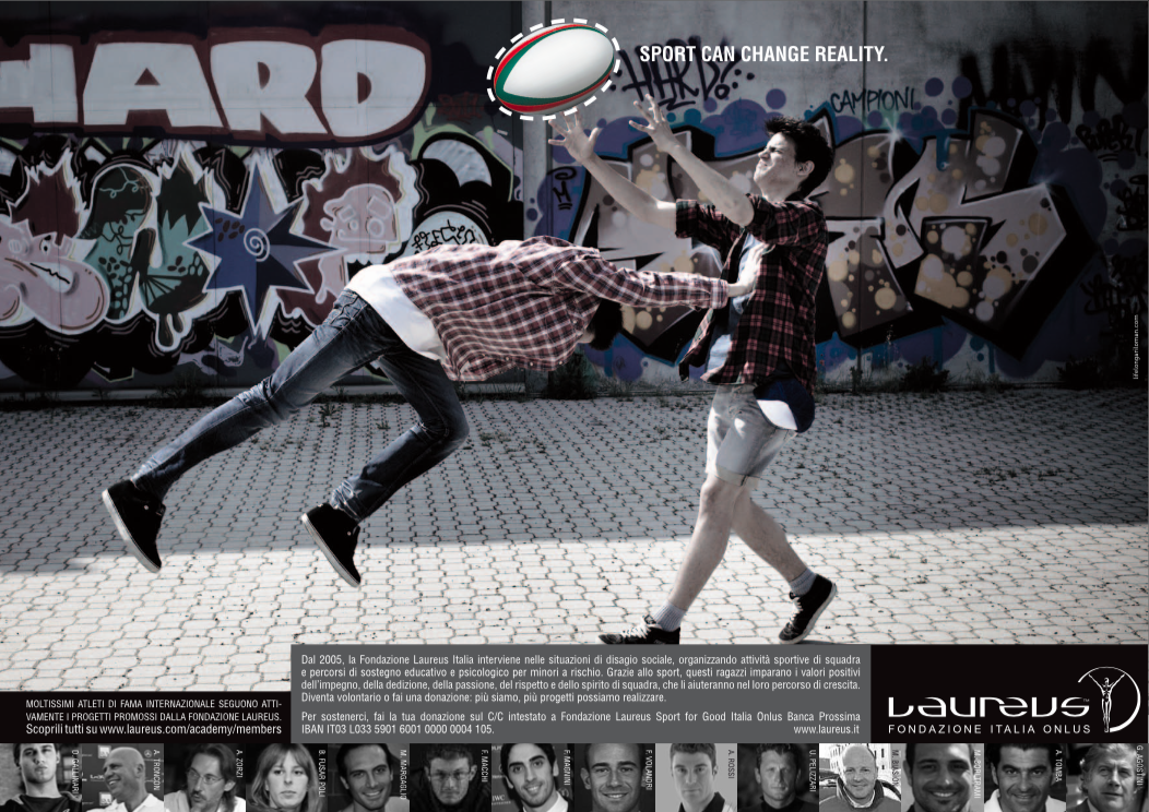 Two young men playing rugby outdoors in front of a graffiti wall, one attempting to catch a rugby ball while the other blocks or intercepts.