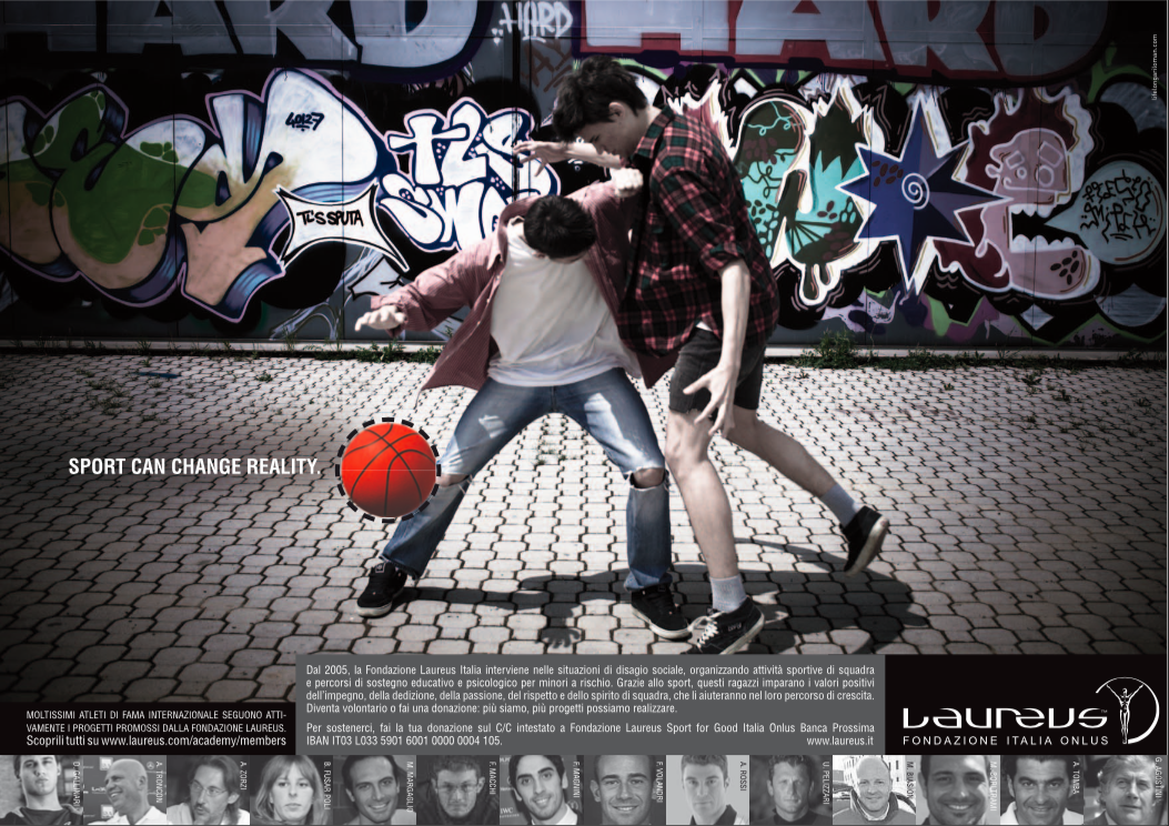 Two young men playing basketball outdoors on a paved surface, with a graffiti wall in the background. One player is wearing a pink jacket and ripped jeans, the other a plaid shirt and shorts.