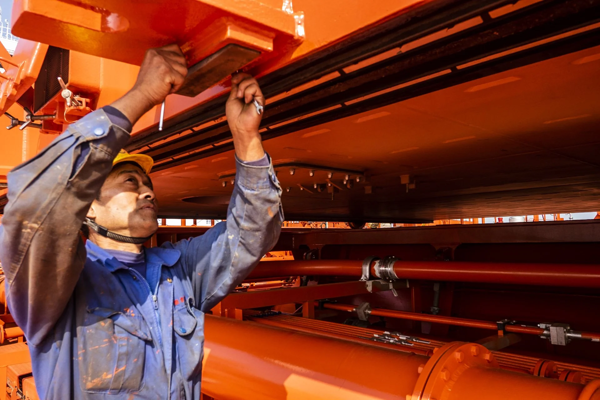 A worker in a blue coverall holds a large orange metal piece under a machine part in an industrial setting with orange machinery.
