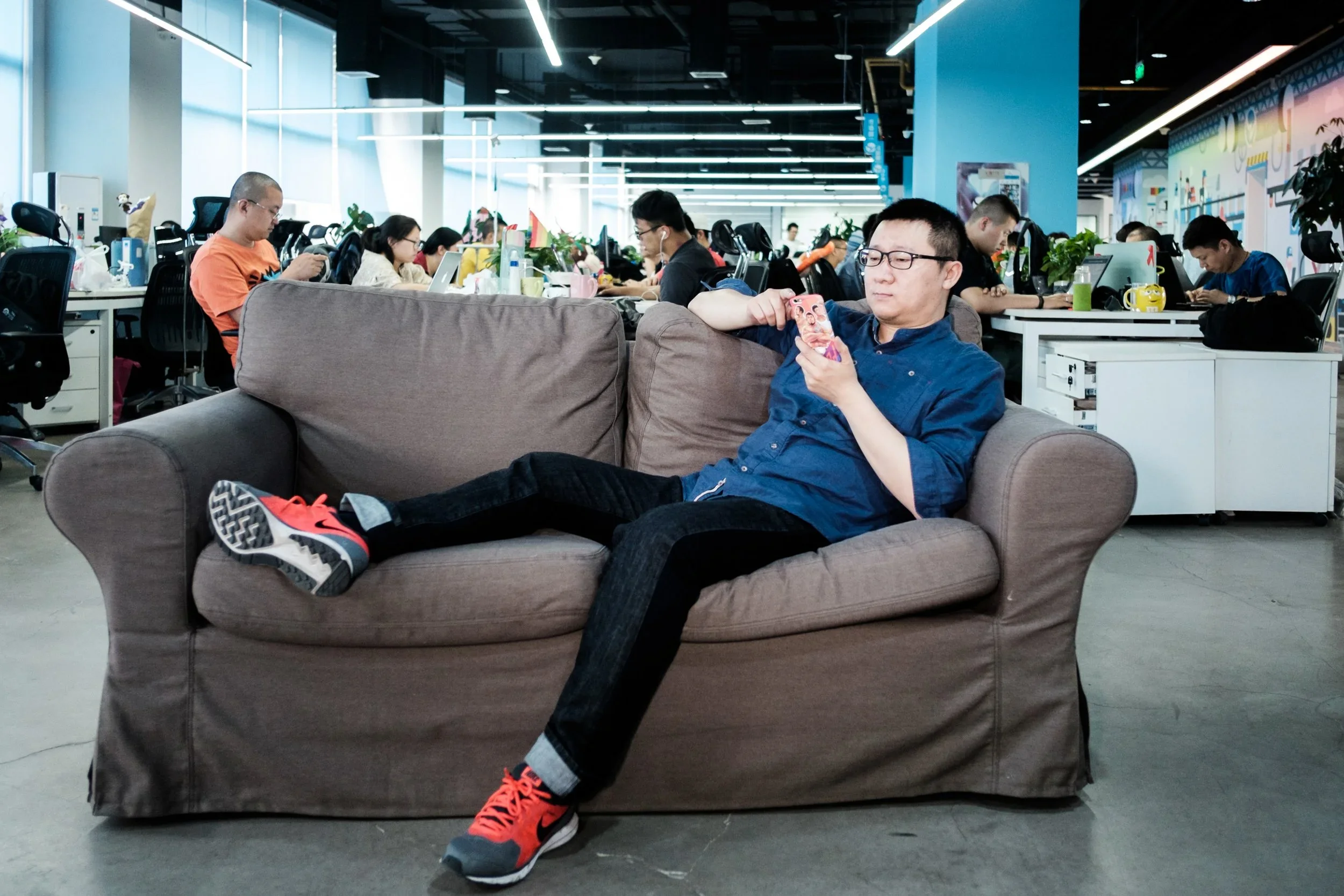 A woman relaxing on a gray sofa in a modern office space, looking at her phone. People are working at desks in the background with computers and various items, illuminated by bright overhead lighting.