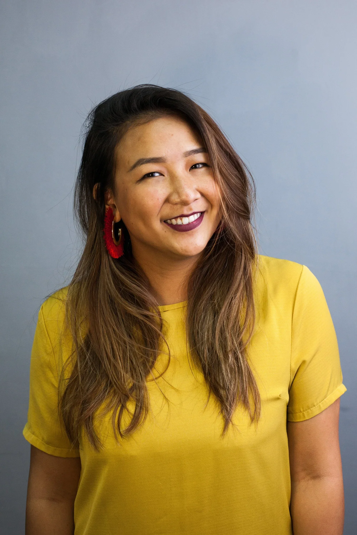 A woman with long, wavy hair and wearing a yellow top is smiling at the camera. She has dark lipstick, hoop earrings with red accents, and is standing against a plain blue background.