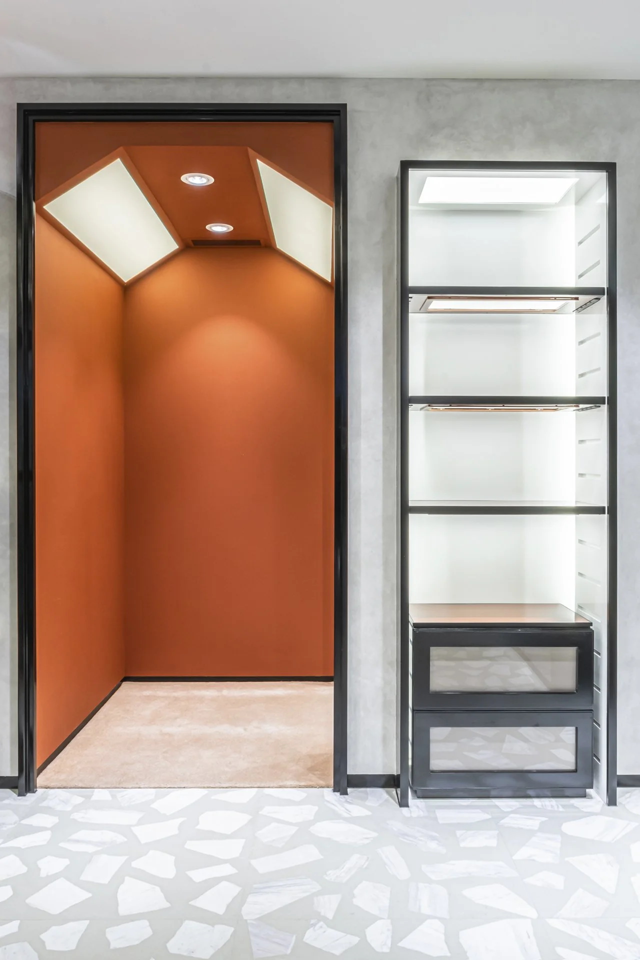 An elevator with orange interior walls and ceiling, and a separate glass-fronted display case or storage unit beside it, set against a light-colored wall.