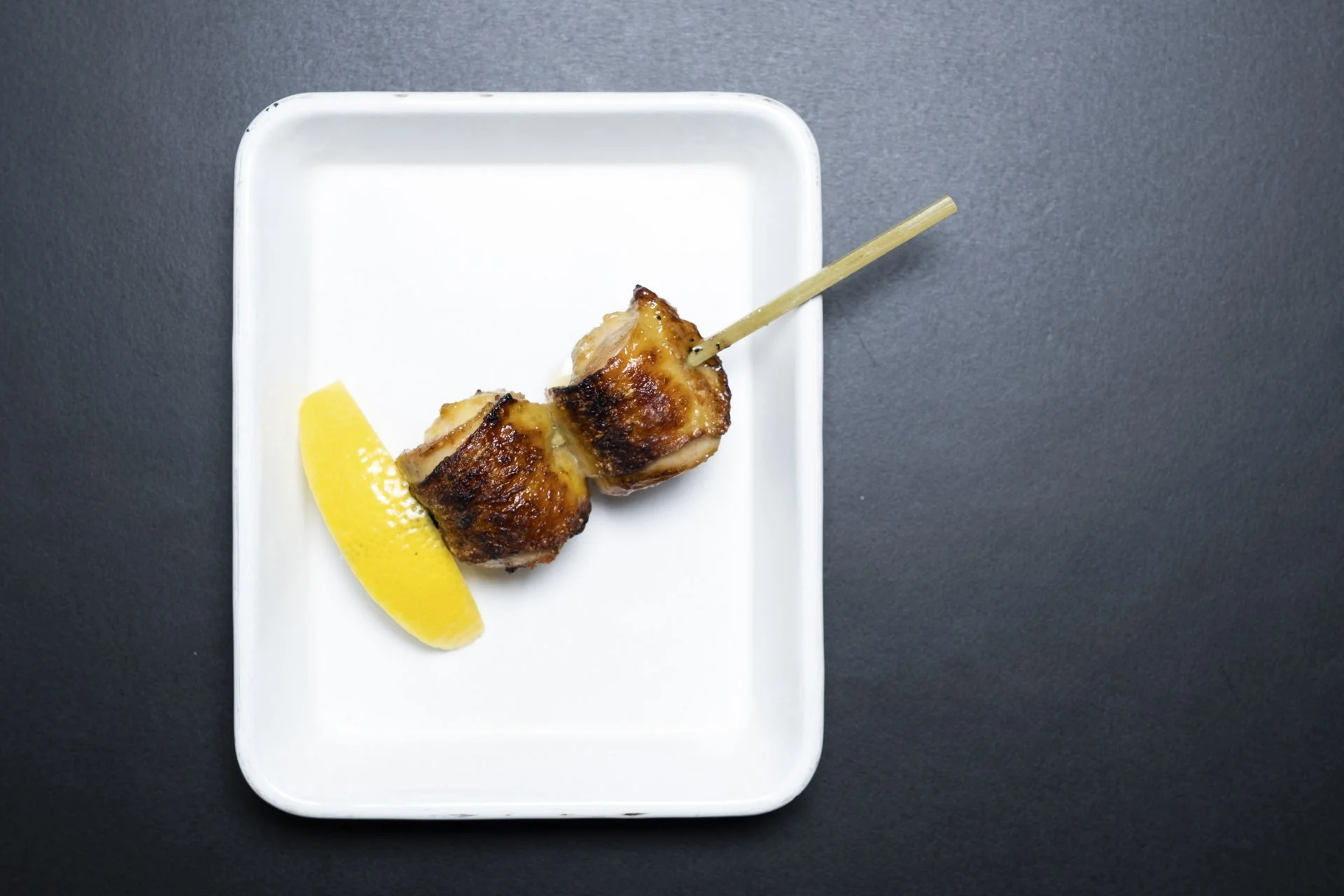 Two grilled chicken skewers on a white rectangular plate, garnished with a lemon wedge, on a dark surface.