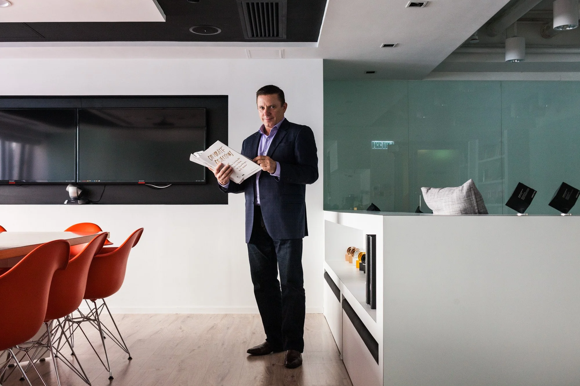 A man dressed in a dark suit and purple shirt standing in a modern, minimalistic office or conference room, holding some papers, with a large black screen on the wall behind him and orange chairs around a wooden table in the foreground.